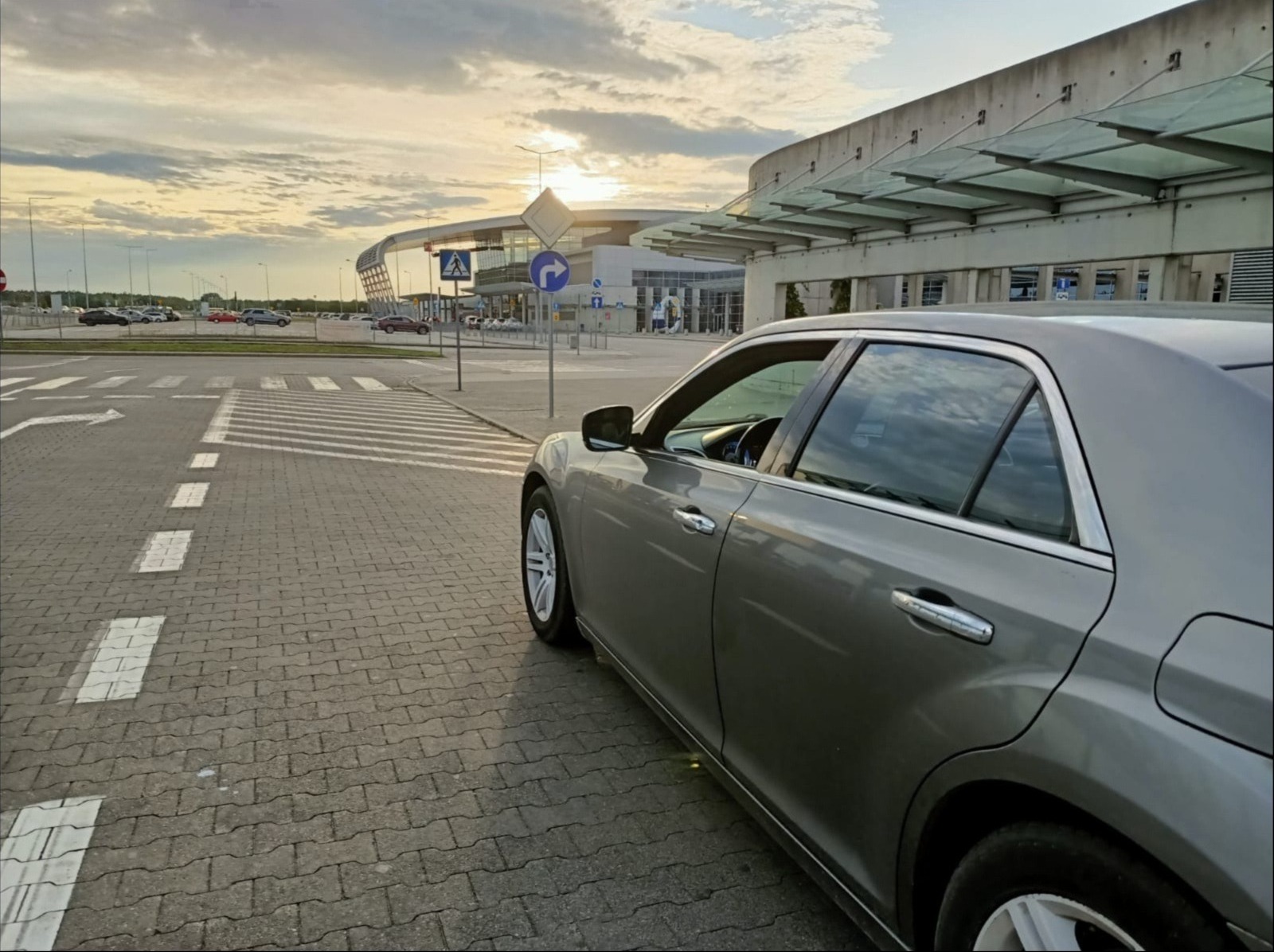 Szary samochód osobowy zaparkowany na kostce brukowej przed nowoczesnym budynkiem lotniska w promieniach zachodzącego słońca, widoczny znak przejścia dla pieszych i oznakowanie poziome.