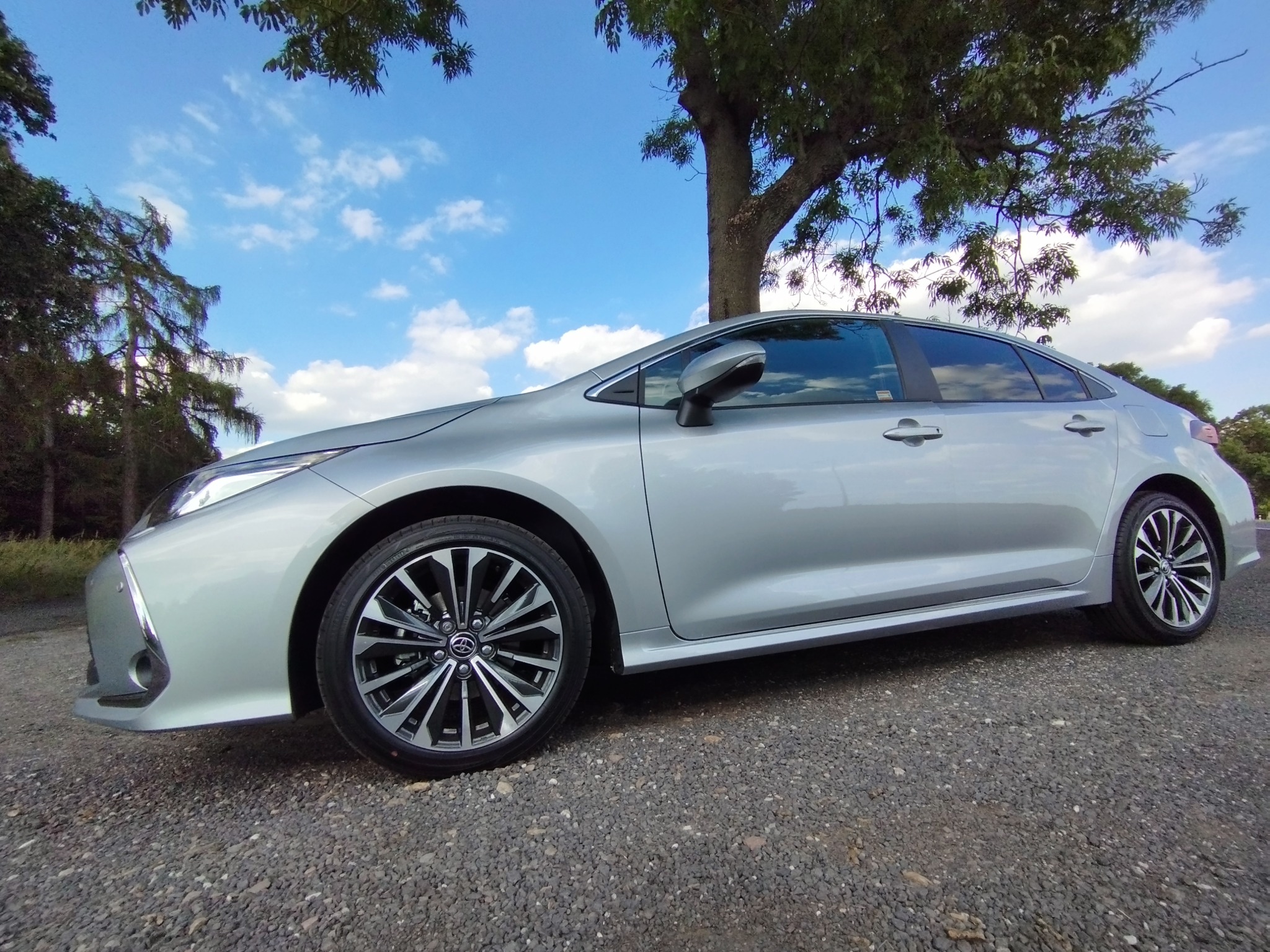 Srebrny sedan Toyota Corolla na żwirowym podłożu z widocznym błękitnym niebem i drzewami w tle.