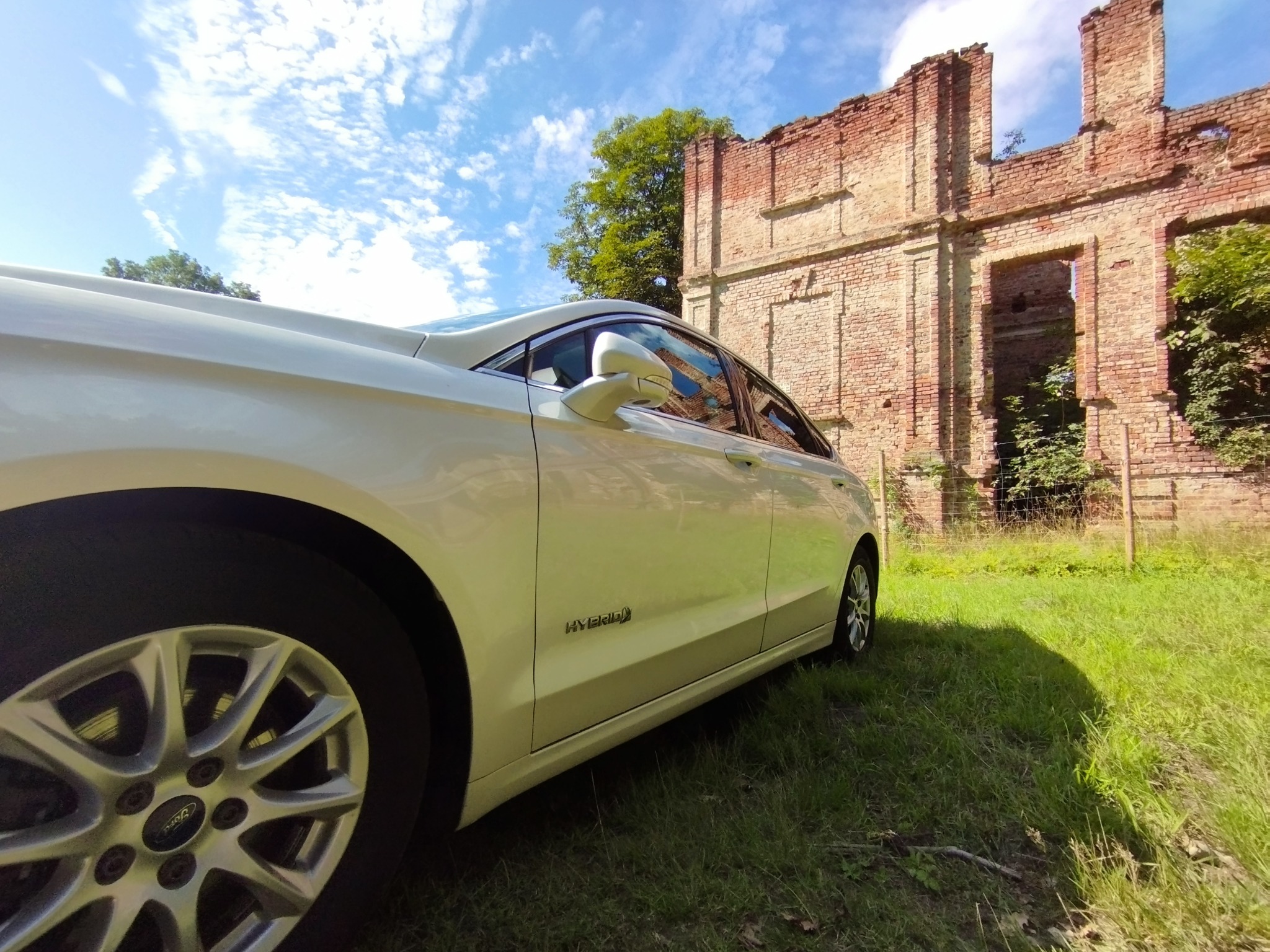 Biały samochód typu sedan, z napisem 'Hybrid' na boku, zaparkowany na trawie przed ruinami ceglanego budynku w słoneczny dzień.