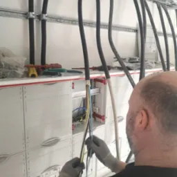 Elektryk w rękawicach łączy przewody w skrzynce rozdzielczej; widoczne czarne kable prowadzone w korytkach kablowych pod sufitem, narzędzia na górnej półce skrzynki.