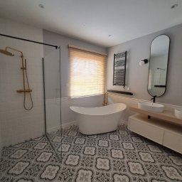 Tamaka Investments Sp. z o. o. - Nowoczesna łazienka z wolnostojącą białą wanną, złotą armaturą, kabiną prysznicową ze złotym deszczownicą, dwoma owalnymi umywalkami na drewnianym blacie, grzejnikiem drabinkowym i wzorzystą podłogą.