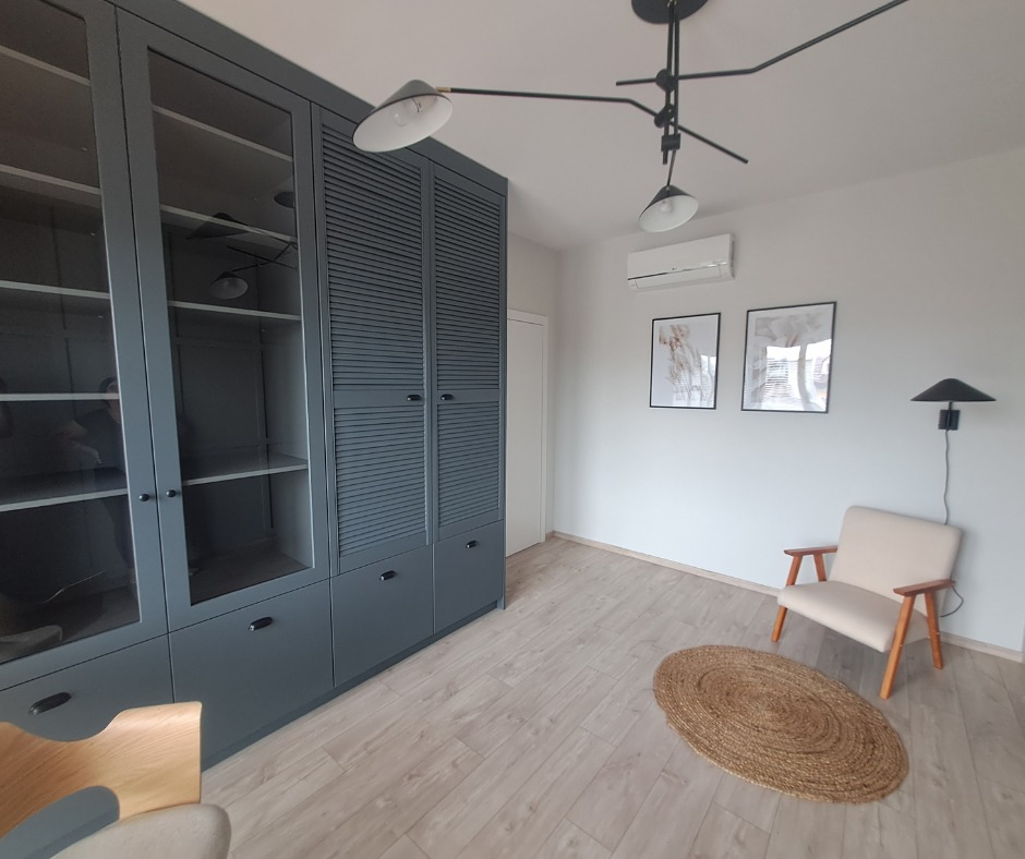 Nowoczesny pokój z szarą zabudową meblową z lamelami i przeszkleniami, drewnianą podłogą, beżowym fotelem i okrągłym dywanem z juty. Na ścianie wiszą grafiki w czarnych ramkach i lampa ścienna.