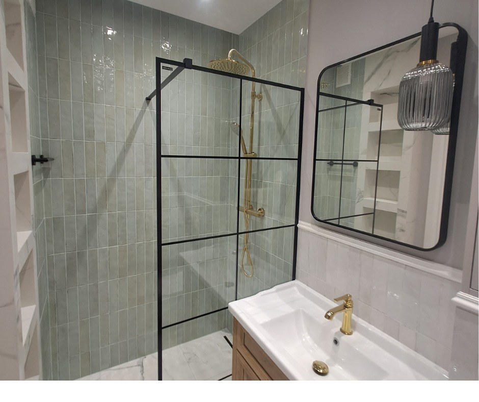 Nowoczesna łazienka z kabiną prysznicową typu walk-in z czarnymi profilami, zielonymi płytkami na ścianach, złotą armaturą i lustrem w czarnej ramie, z widoczną ozdobną lampą wiszącą.