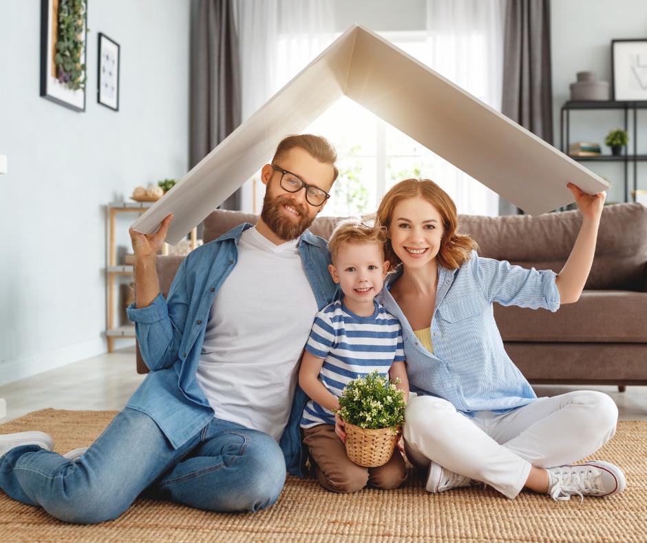 Uśmiechnięta rodzina z dzieckiem siedzi na podłodze, trzymając nad głowami białą płytę imitującą dach domu, z kwiatami w koszyku na pierwszym planie.