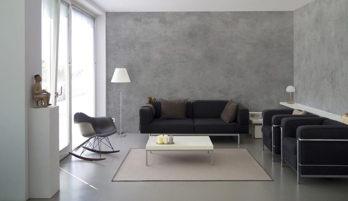 Salon z szarą ścianą wykończoną w stylu betonu architektonicznego, czarną sofą i fotelami na metalowych nogach, biały stolik kawowy z trzema świecami, lampa podłogowa i rzeźba siedzącego mężczyzny...