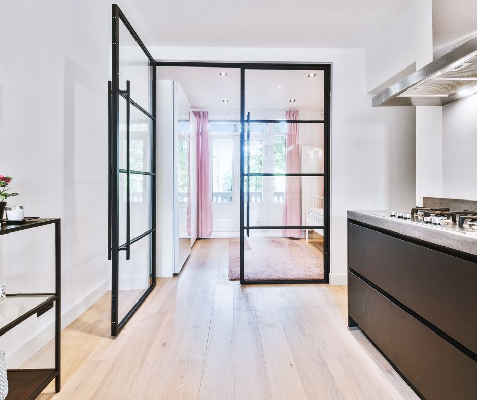 Nowoczesne, przeszklone drzwi z czarnymi szprosami oddzielają kuchnię od pokoju z różowymi zasłonami i dywanem, widoczne jasne panele podłogowe.