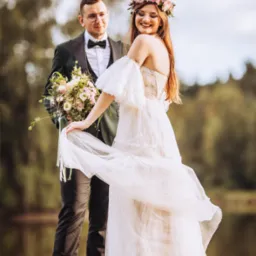 Uśmiechnięta panna młoda w zwiewnej sukni z kwiatowym wiankiem na głowie, trzymająca brzeg sukni, obok pan młody w ciemnozielonym garniturze z bukietem, na drewnianym pomoście nad jeziorem.