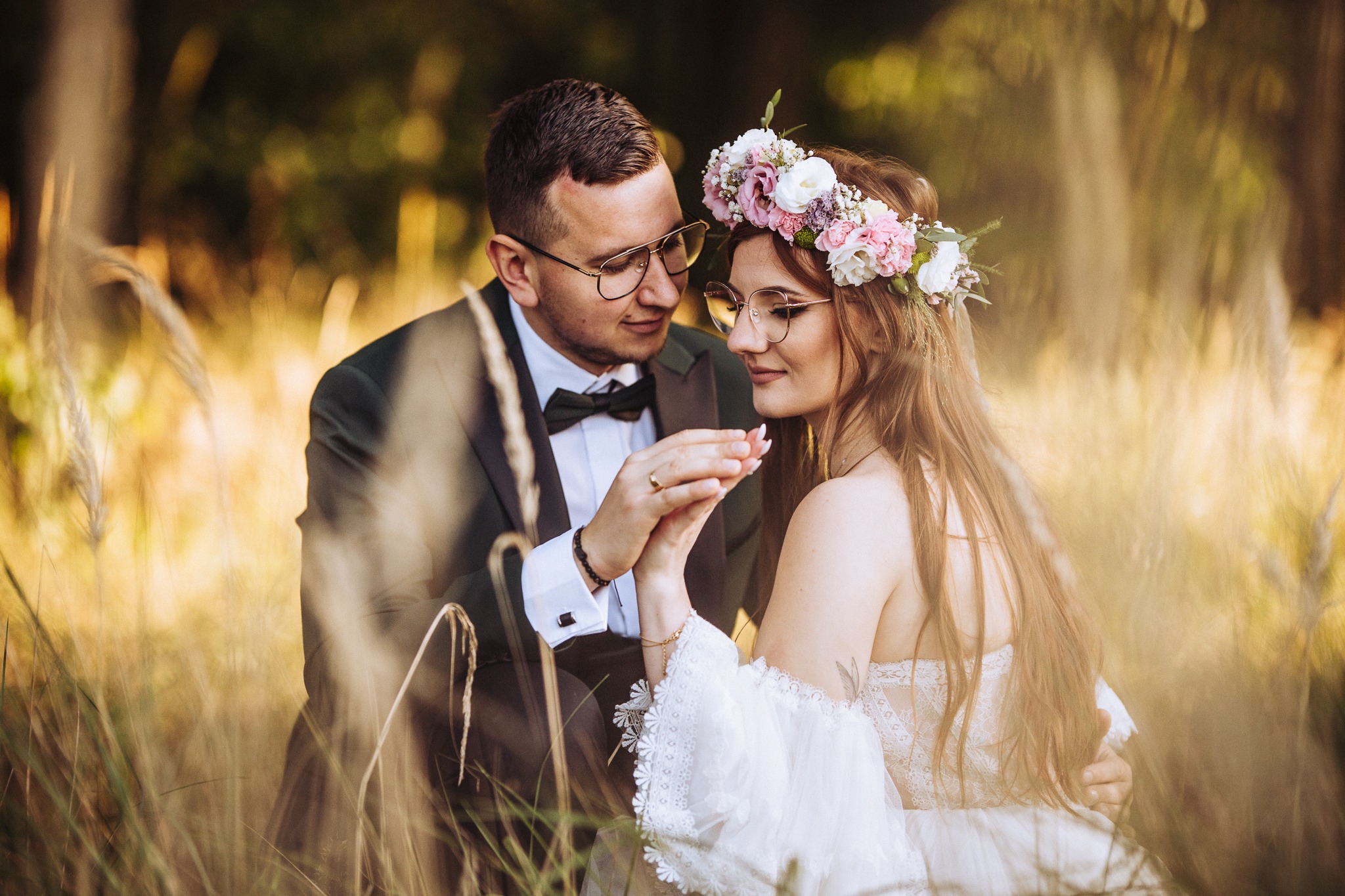 Para młoda w plenerze, panna młoda z wiankiem z różowych i białych kwiatów, pan młody w ciemnozielonym smokingu i okularach, trzymają się za ręce na tle wysokiej trawy.