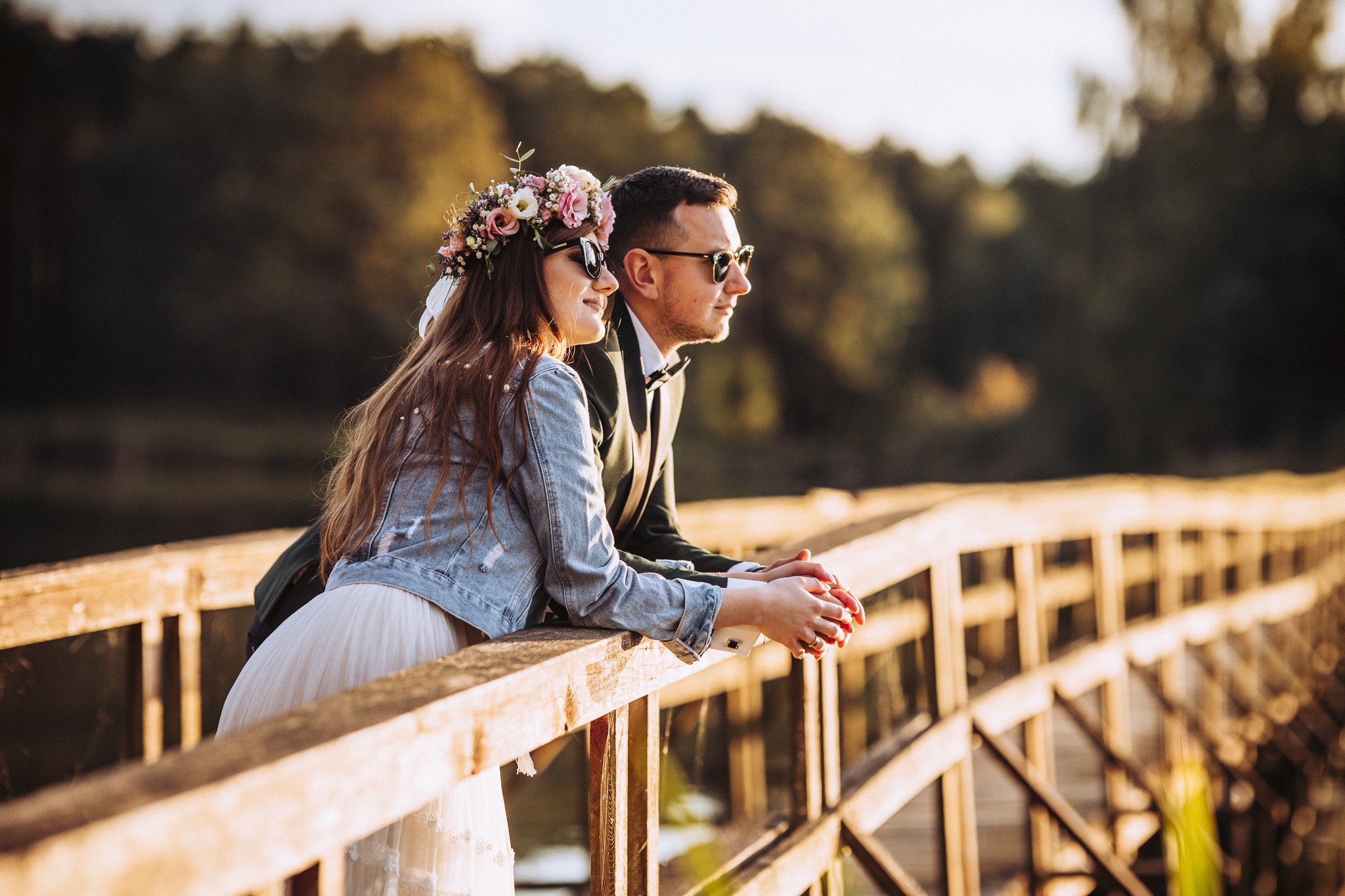 Para młoda w okularach przeciwsłonecznych, oparta o drewnianą barierkę mostu, panna młoda z wiankiem z różowych i białych kwiatów na głowie, w denimowej kurtce i tiulowej sukience, w tle zieleń drzew.