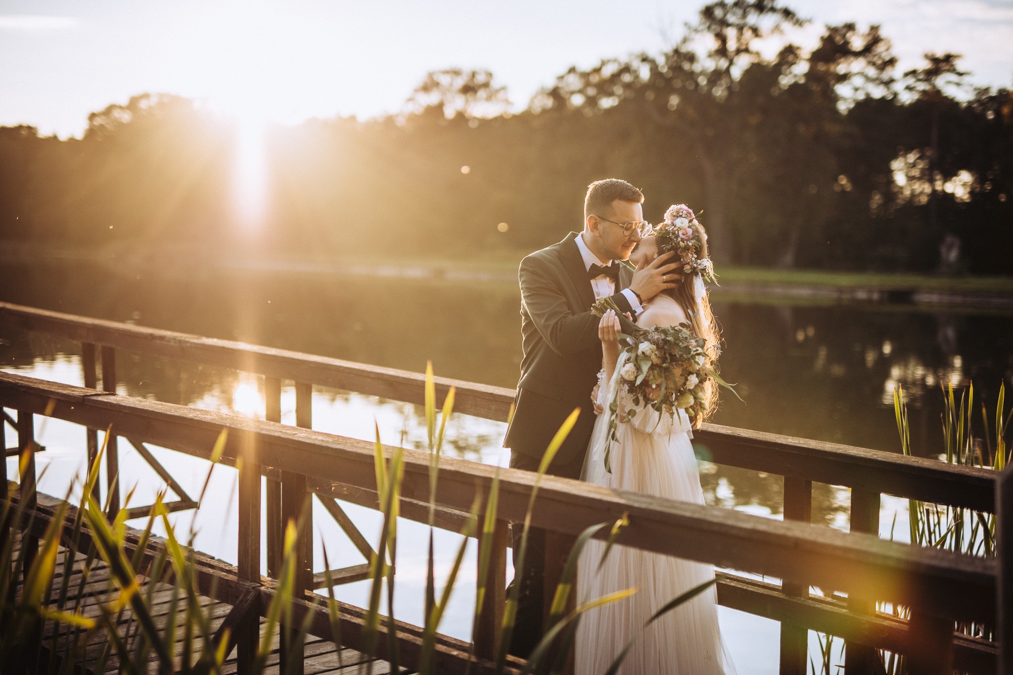 Para młoda w czułym uścisku na drewnianym pomoście nad jeziorem w promieniach zachodzącego słońca. Panna młoda w sukni z kwiatowym wiankiem, pan młody w ciemnym garniturze z muszką.