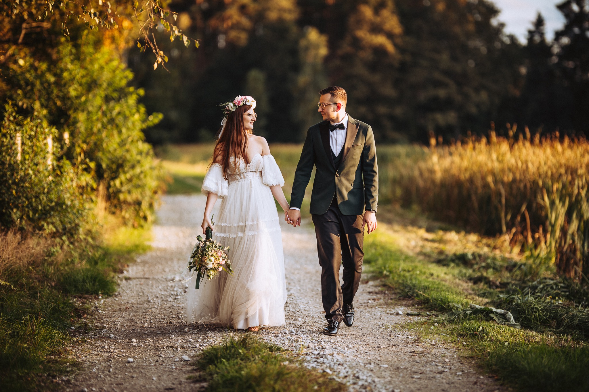 Para młoda spaceruje po polnej drodze w promieniach zachodzącego słońca; panna młoda w zwiewnej sukni z kwiatowym wiankiem i bukietem, pan młody w ciemnozielonym smokingu i okularach.