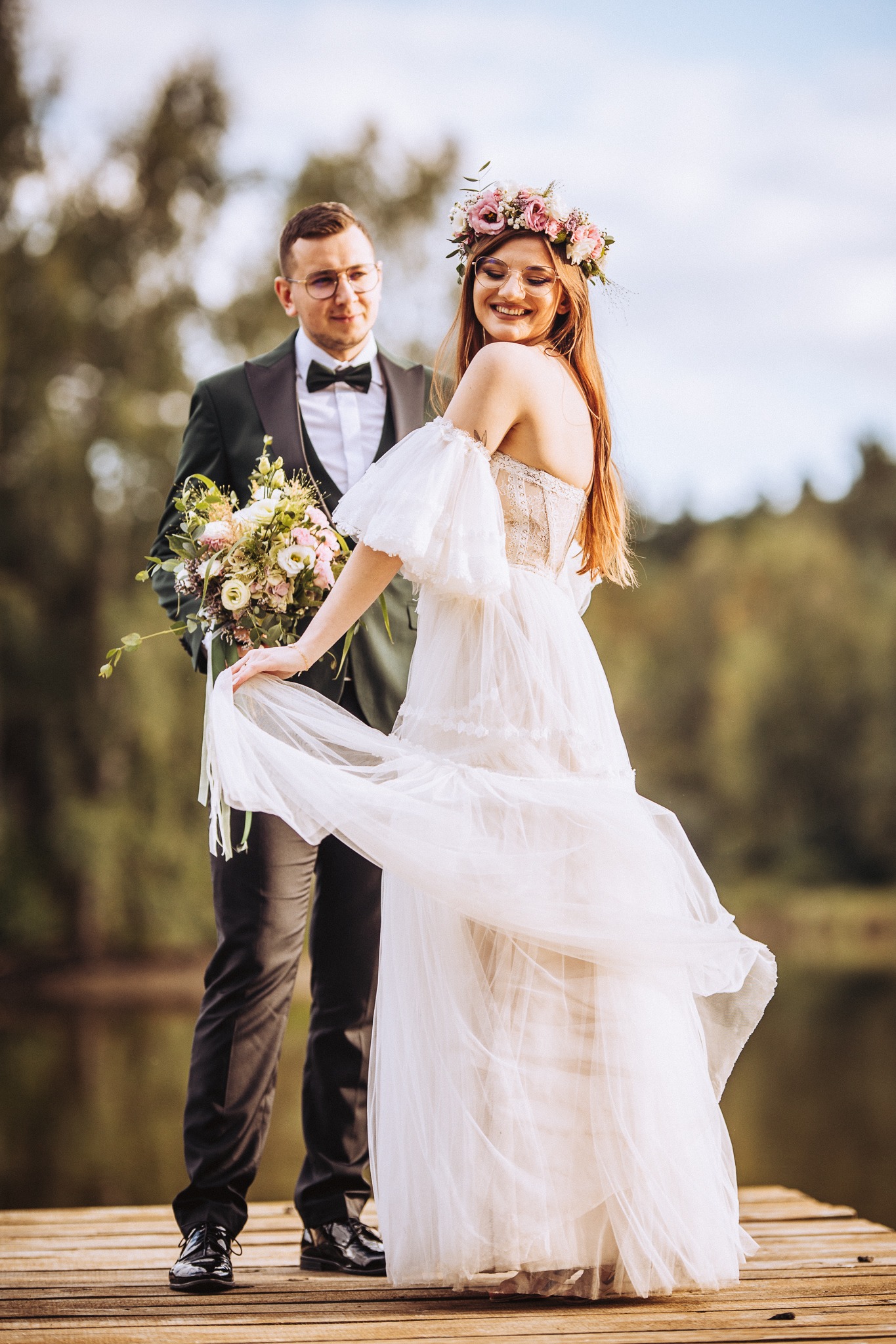 Uśmiechnięta panna młoda w zwiewnej sukni z kwiatowym wiankiem na głowie, trzymająca brzeg sukni, obok pan młody w ciemnozielonym garniturze z bukietem, na drewnianym pomoście nad jeziorem.