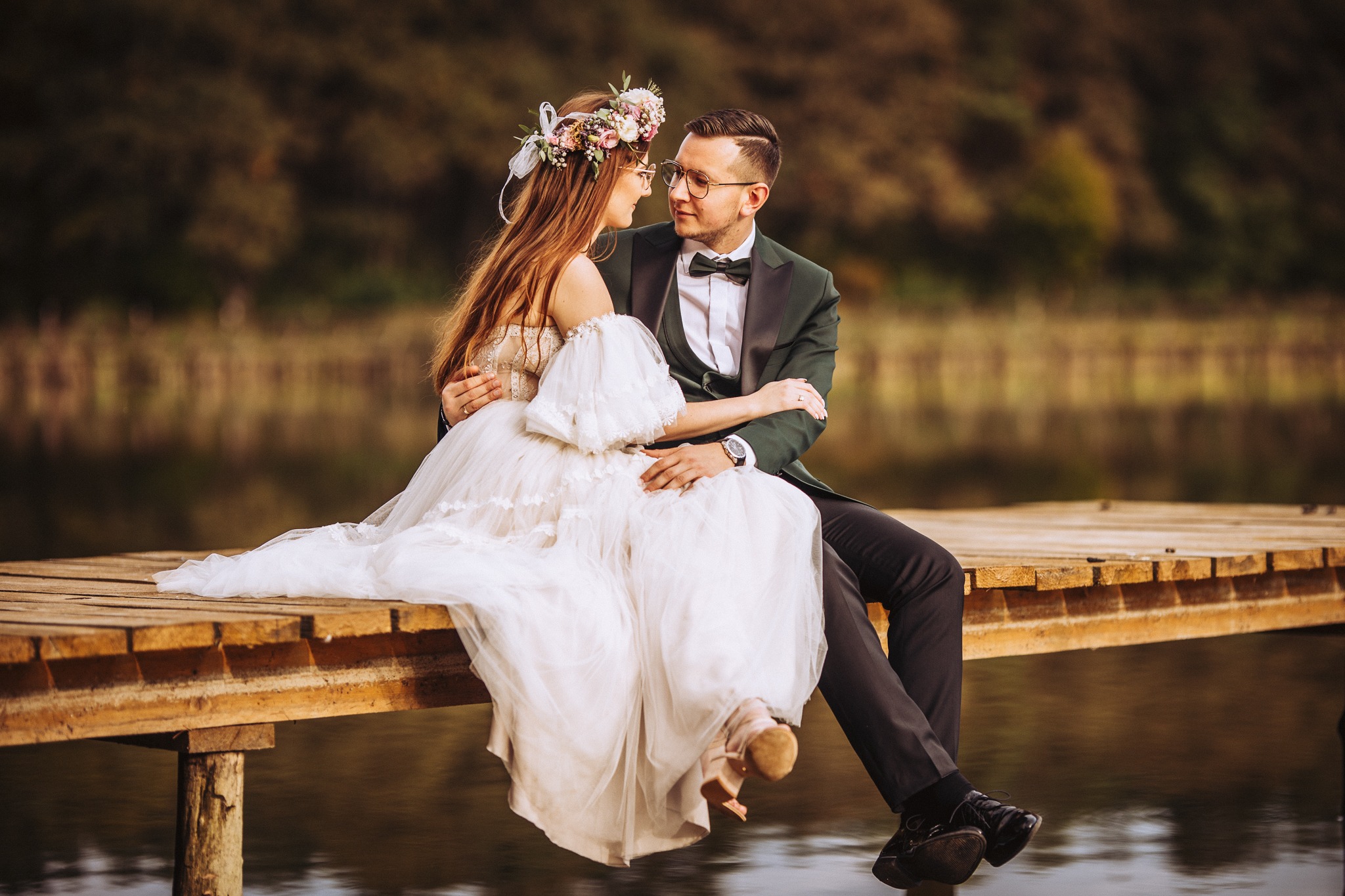 Para młoda siedząca na drewnianym pomoście nad wodą, panna młoda w sukni z bufiastymi rękawami i wiankiem z kwiatów, pan młody w ciemnozielonym smokingu z muchą, oboje patrzą na siebie z czułością.