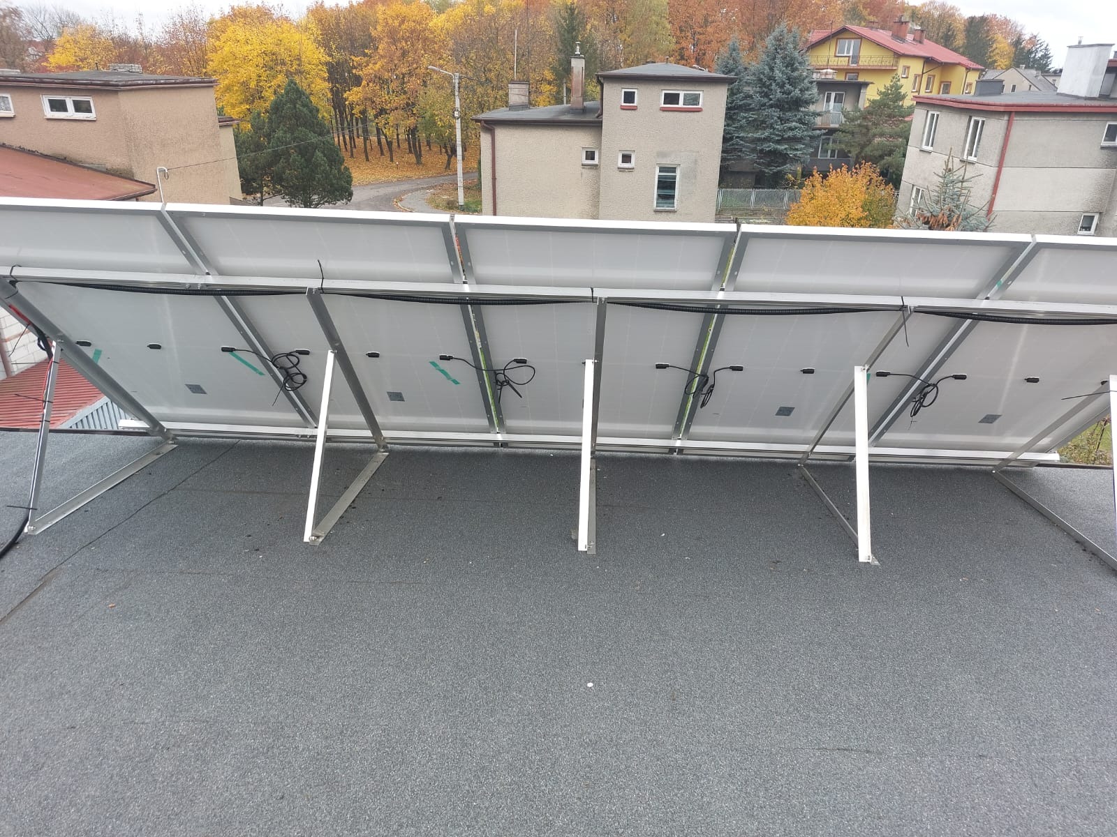 Widok od tyłu na panele słoneczne zamontowane na płaskim dachu, z widocznymi kablami i elementami mocującymi, w tle jesienny krajobraz z budynkami.