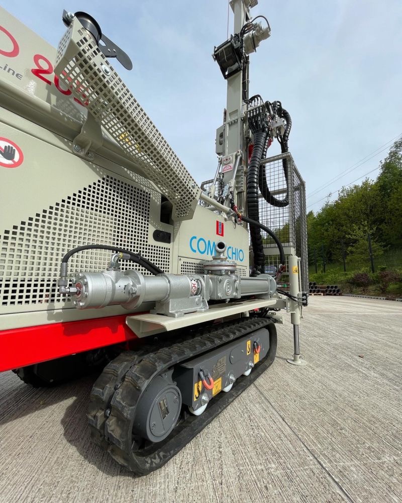 Mobilna wiertnica Comacchio na gąsienicach, widok z dołu, z wysuniętą podporą stabilizującą, na betonowej nawierzchni, z zielenią w tle.