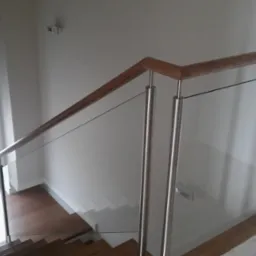 Szklana balustrada schodowa z drewnianą poręczą i metalowymi słupkami, widok z góry na schody z drewnianymi stopniami.