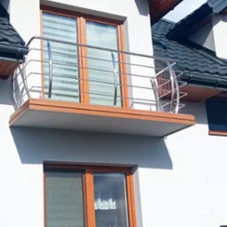 Balkon z poziomymi, metalowymi barierkami na tle białej elewacji domu z oknami w drewnianych ramach.