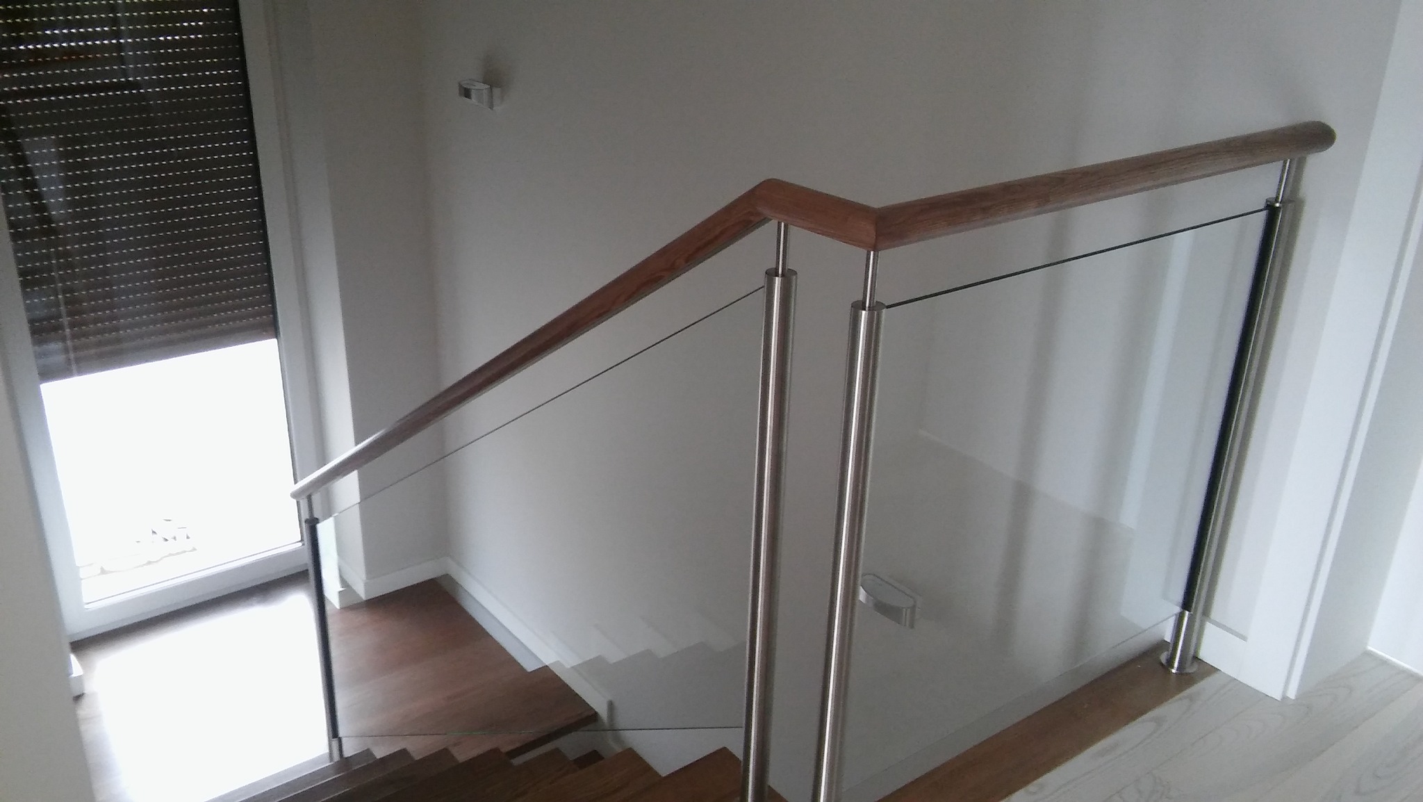 Szklana balustrada schodowa z drewnianą poręczą i metalowymi słupkami, widok z góry na schody z drewnianymi stopniami.