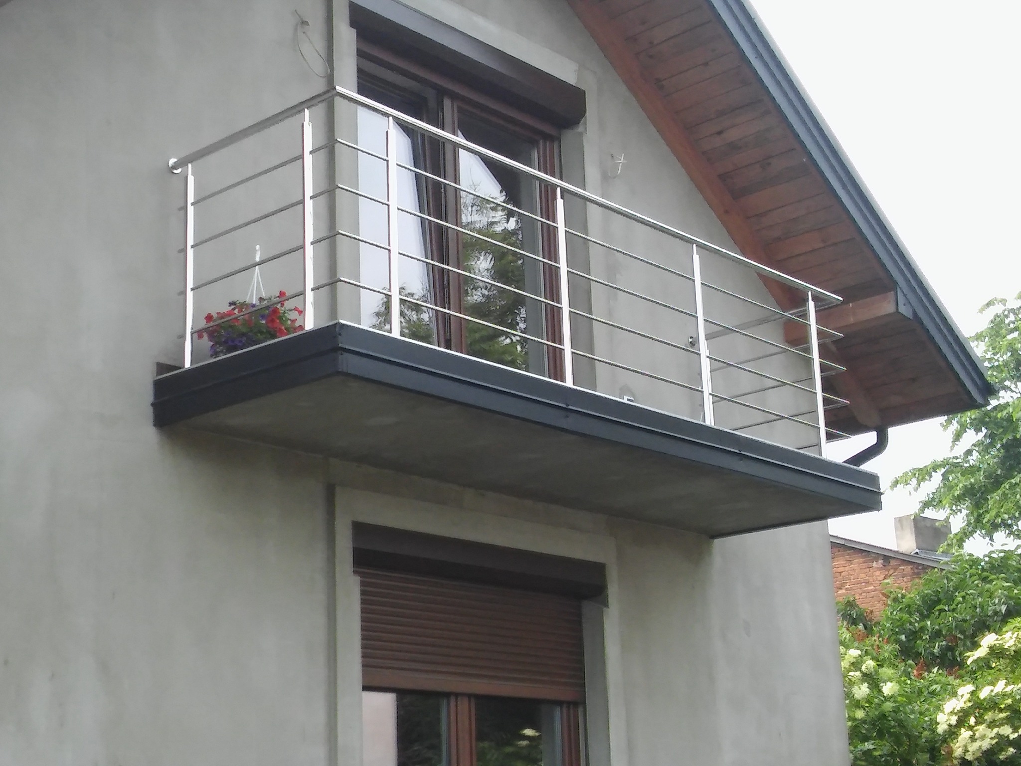 Balkon z nowoczesną, metalową barierką z poziomych prętów, z doniczką z kwiatami, widok z dołu na jasną elewację budynku.