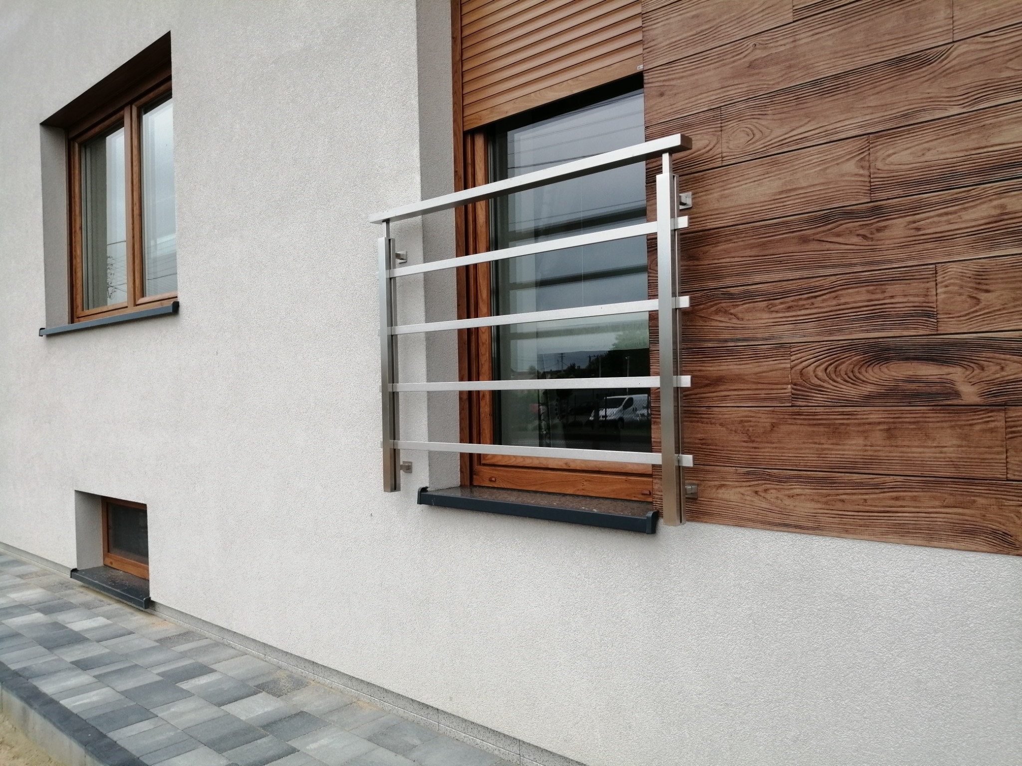 Zewnętrzna, metalowa konstrukcja zabezpieczająca okno, z poziomymi prętami, zamontowana na elewacji budynku z drewnopodobną okładziną i żaluzjami.