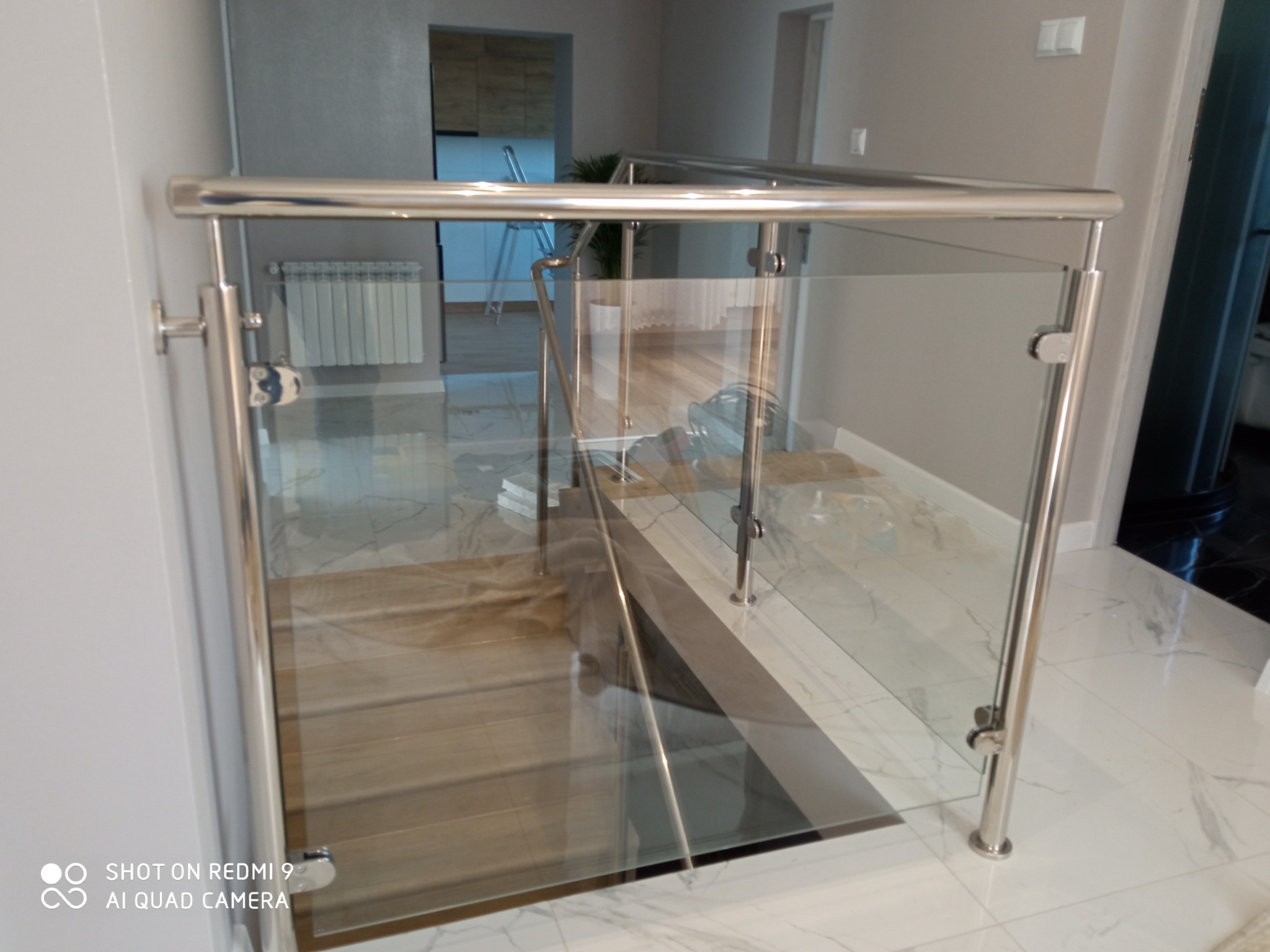 Szklana balustrada ze stalowymi słupkami zamontowana przy schodach wewnętrznych, widoczne mocowania szkła do słupków, marmurowa podłoga.