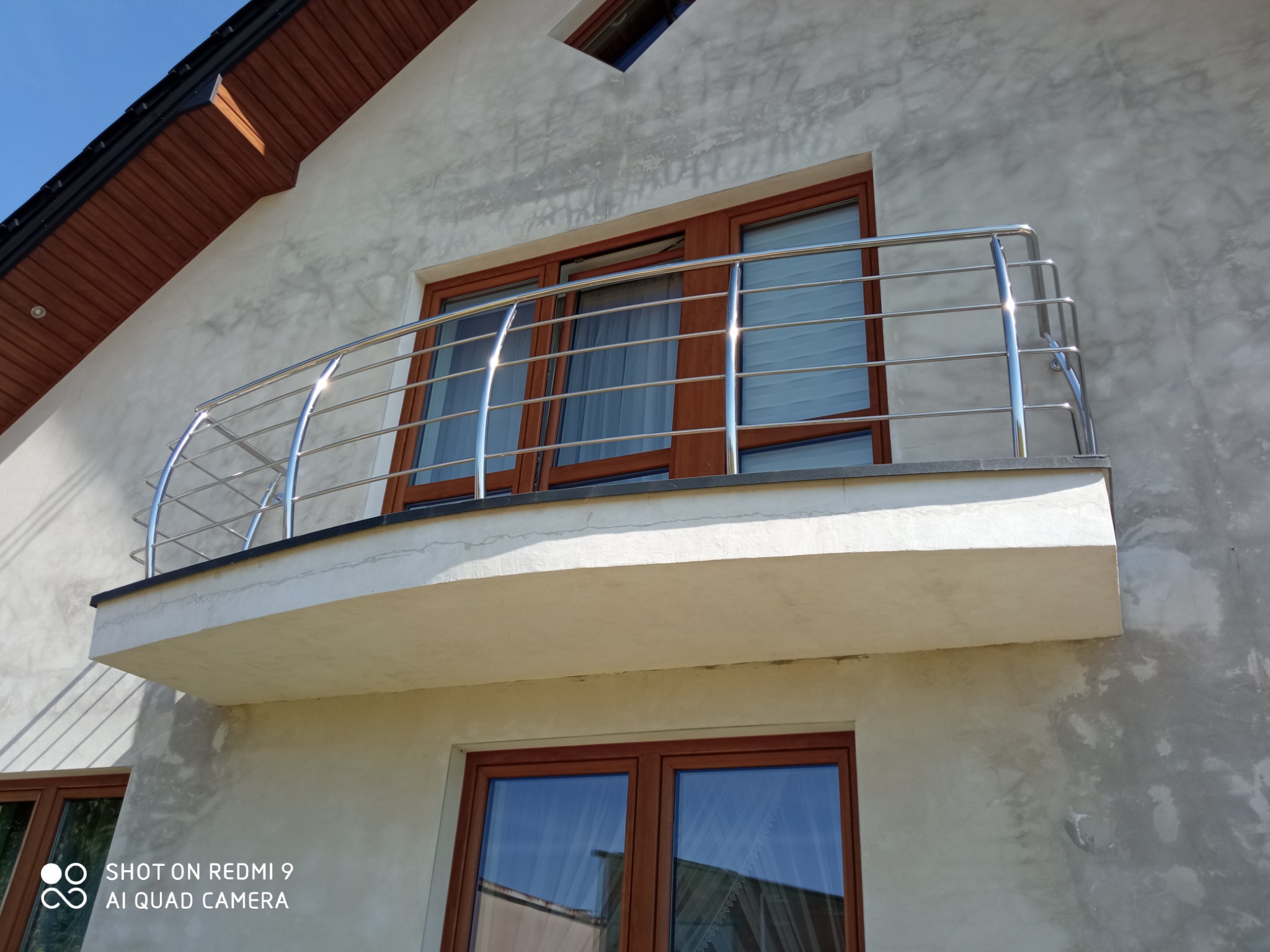 Balkon z zaokrągloną, metalową konstrukcją zabezpieczającą, widok z dołu na tle surowej elewacji budynku.