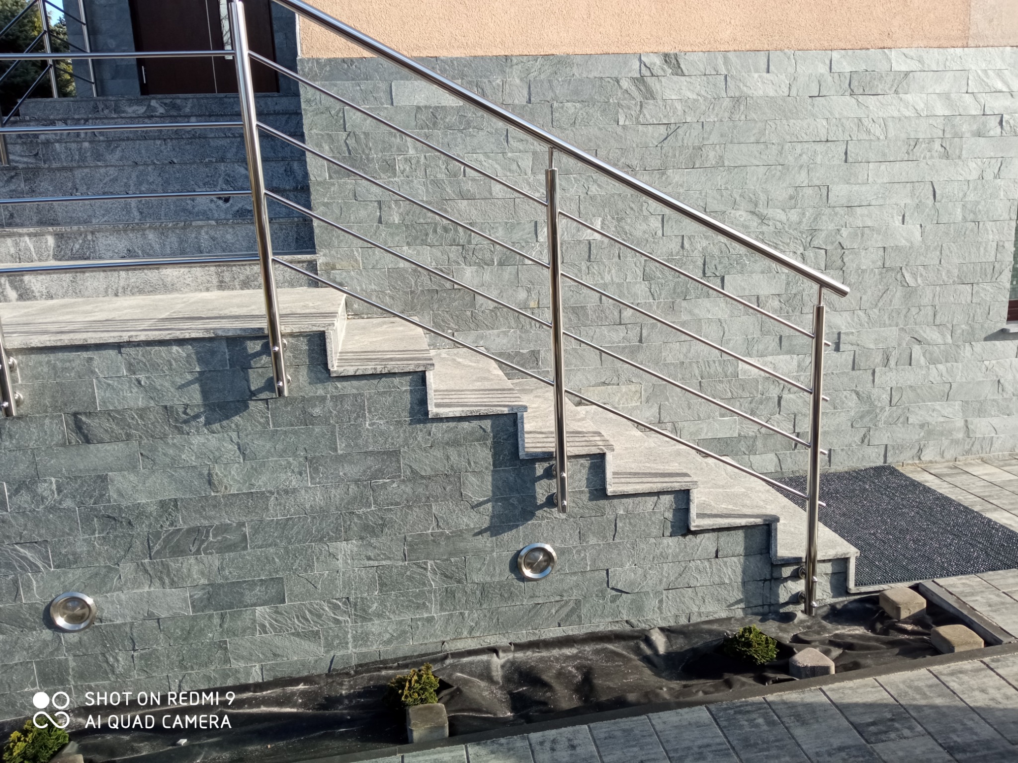 Zewnętrzne schody wyłożone szarą kamienną okładziną z zamontowaną balustradą ze stali nierdzewnej o minimalistycznym designie.