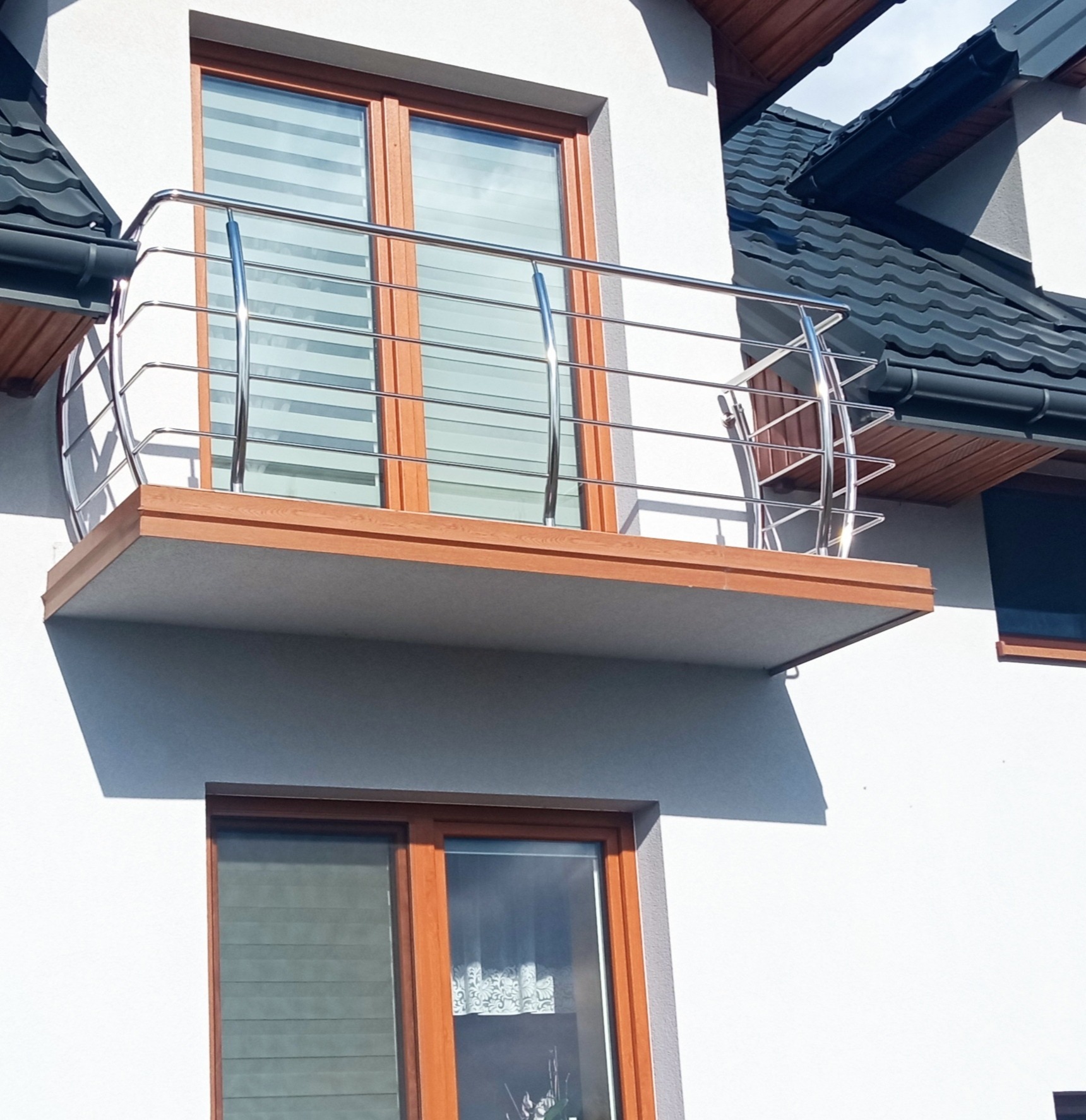 Balkon z poziomymi, metalowymi barierkami na tle białej elewacji domu z oknami w drewnianych ramach.