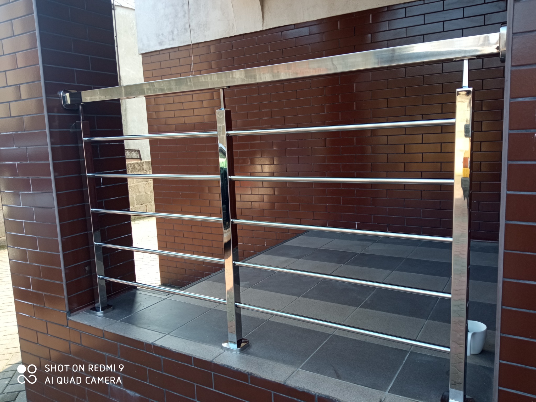 Błyszcząca, chromowana barierka z poziomymi prętami, zamontowana na ceglanym murku, z widocznym fragmentem wyłożonej płytkami podłogi.