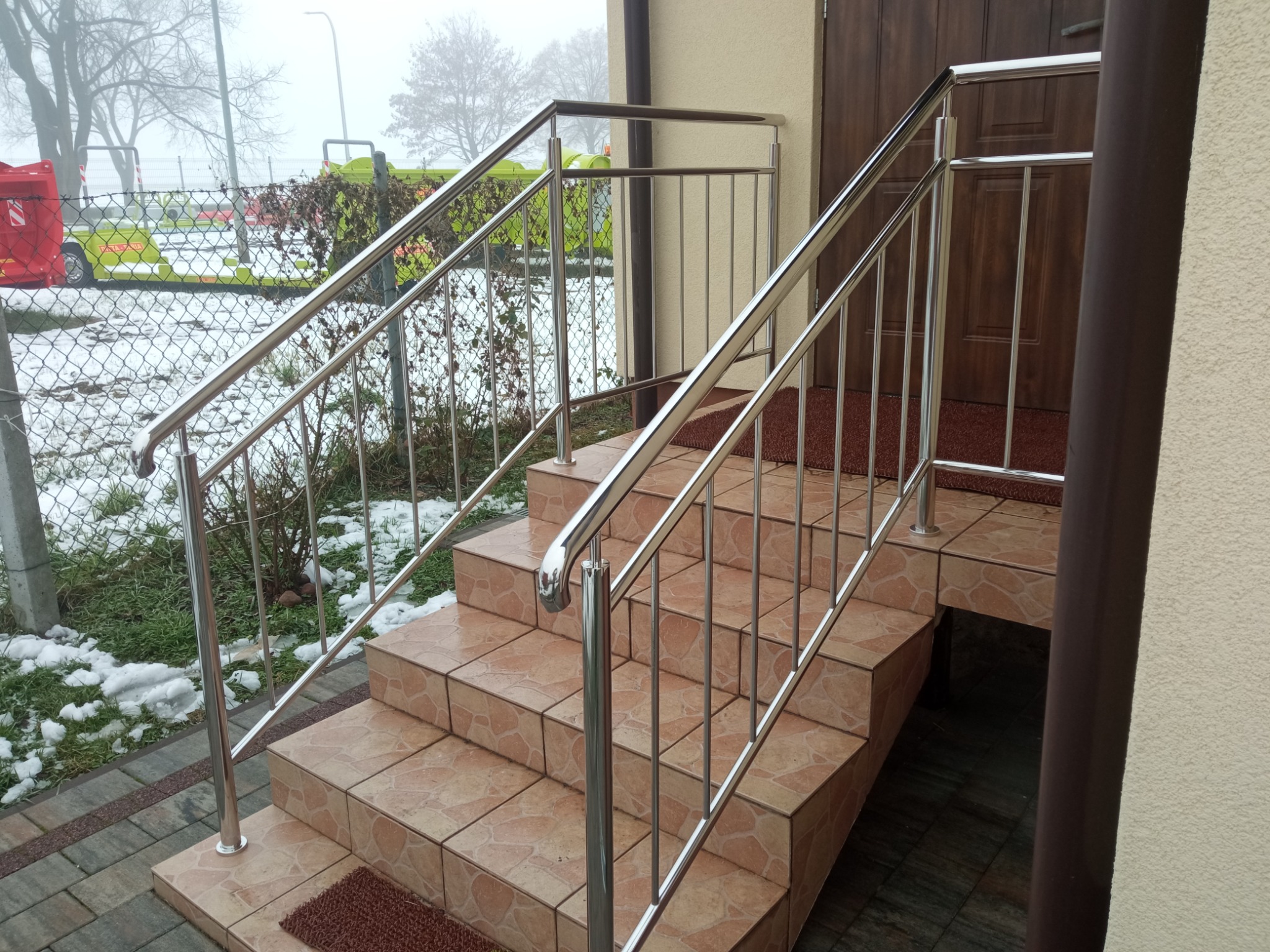 Zewnętrzne schody wejściowe z ceramicznymi płytkami i błyszczącą metalową konstrukcją zabezpieczającą, widok na zewnątrz w pochmurny dzień.