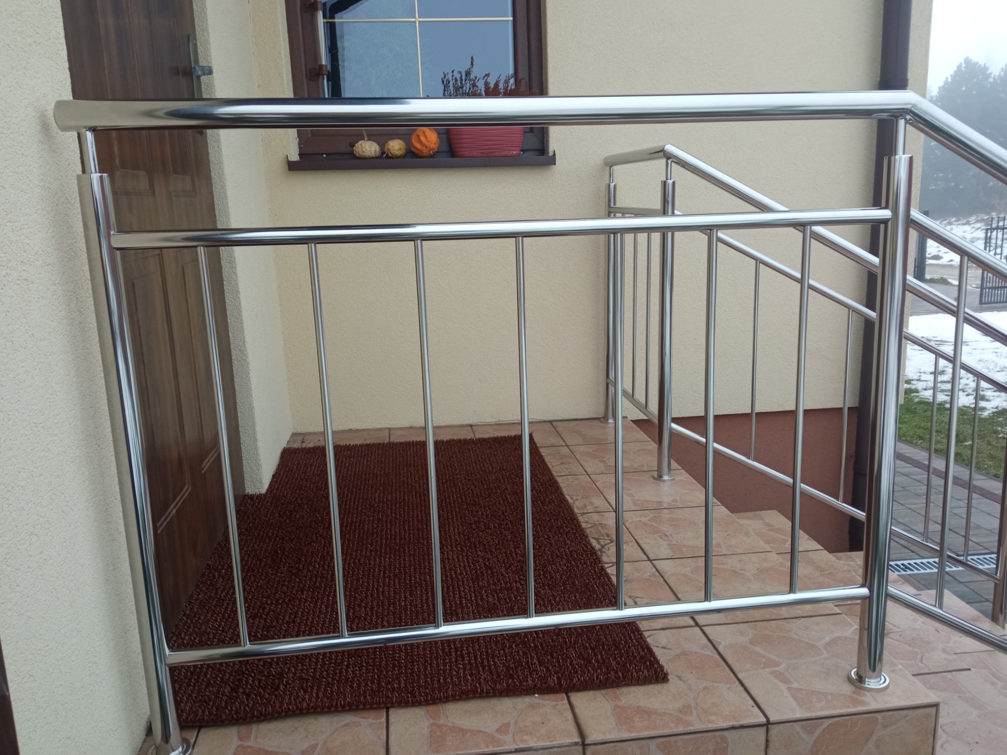 Błyszcząca, metalowa konstrukcja zabezpieczająca wejście do domu, z widoczną brązową wycieraczką i dekoracjami dyniowymi w oknie.