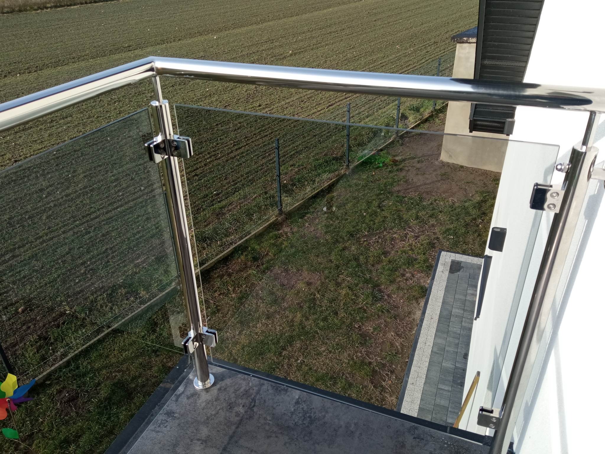 Balkon z nowoczesną szklaną balustradą i polerowaną, metalową ramą, widok z góry na pole i dom sąsiada.