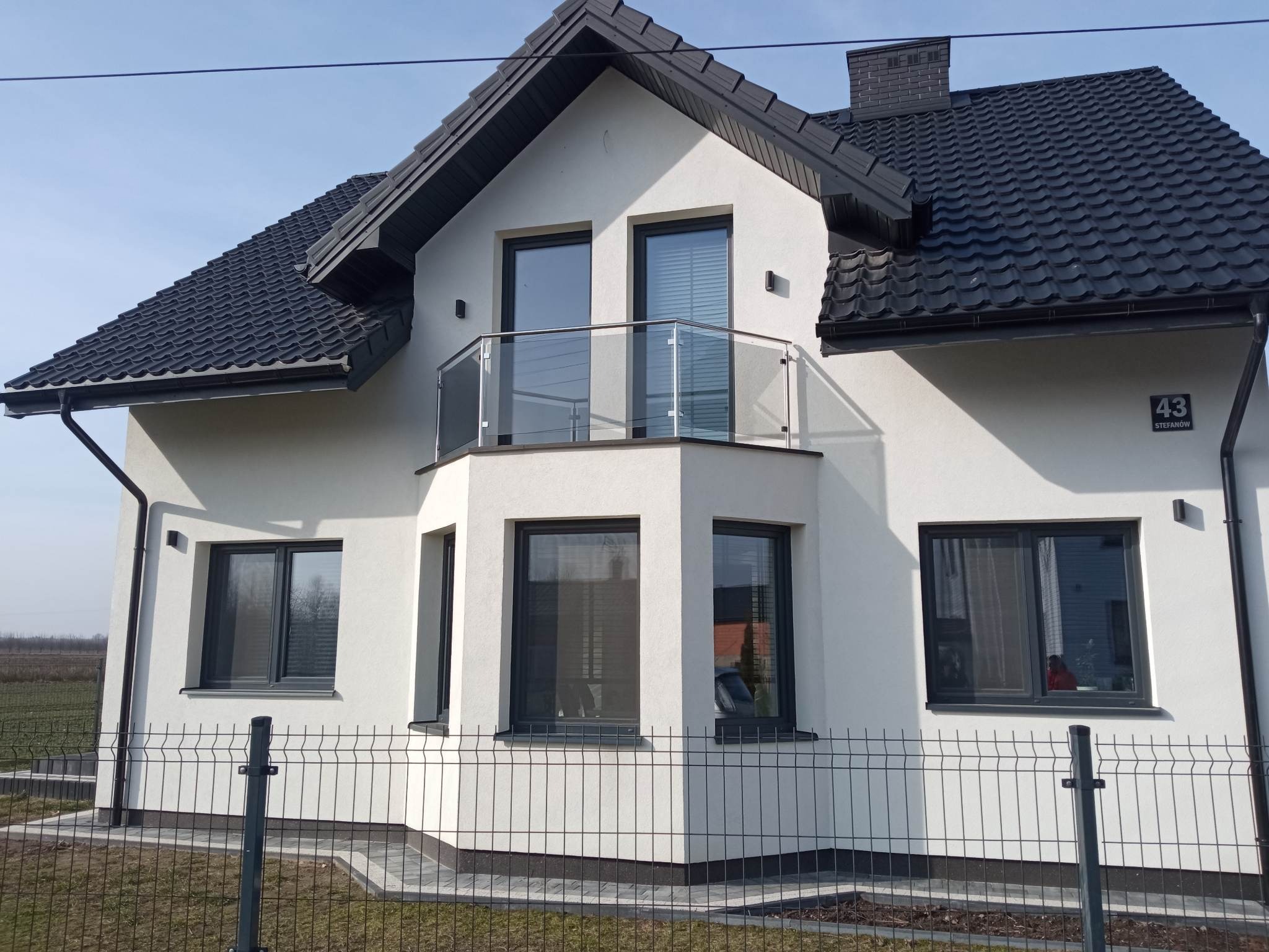 Nowoczesny dom jednorodzinny z balkonem zabezpieczonym szklaną balustradą, widok zza ogrodzenia panelowego.