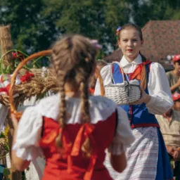 Dwie kobiety w tradycyjnych strojach ludowych, jedna trzyma koszyk, druga wieniec dożynkowy, na tle zieleni.