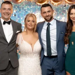 Cztery osoby pozują do zdjęcia na tle błyszczącej ściany: mężczyzna w szarym garniturze z muchą, kobieta w białej sukni z koronki, mężczyzna w niebieskim garniturze z krawatem i kobieta w zielonej...