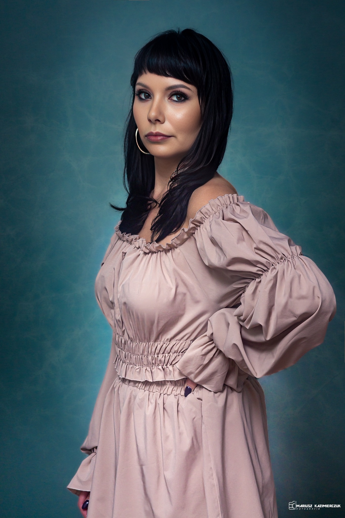 Portret kobiety z ciemnymi włosami i grzywką, w bladoróżowej sukience z odkrytymi ramionami i marszczeniami, pozującej na tle abstrakcyjnej, turkusowej tekstury.