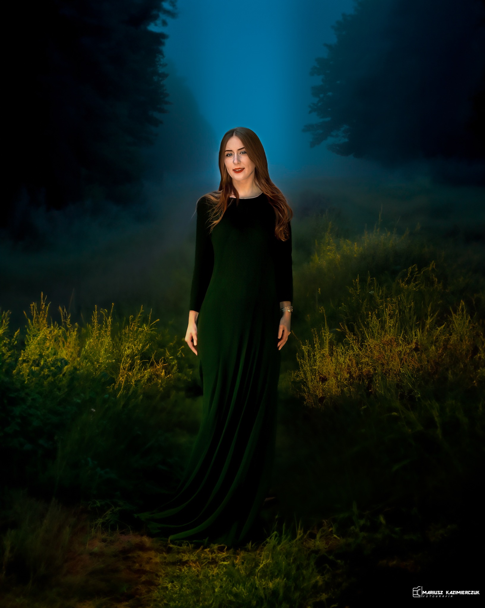 Kobieta w długiej, ciemnozielonej sukni pozuje w leśnej scenerii z mgłą i wysokimi trawami, oświetlona delikatnym światłem.