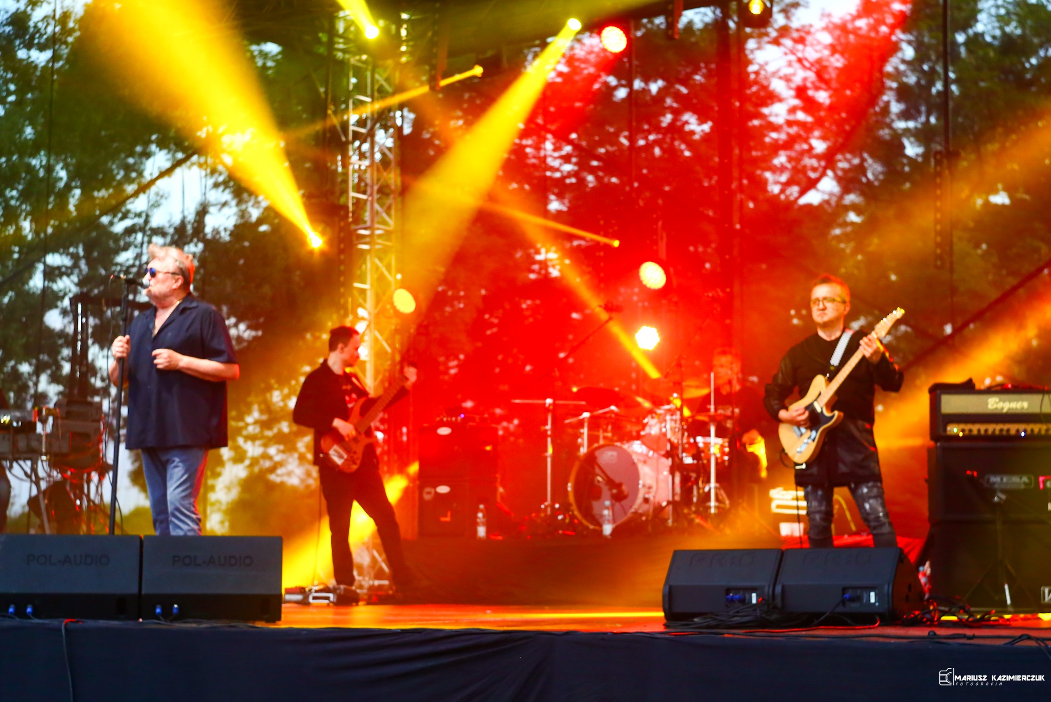 Zespół muzyczny podczas koncertu na scenie, oświetlonej jaskrawymi światłami, z wokalistą w czarnej koszuli i gitarzystami z gitarami elektrycznymi, perkusja w tle.