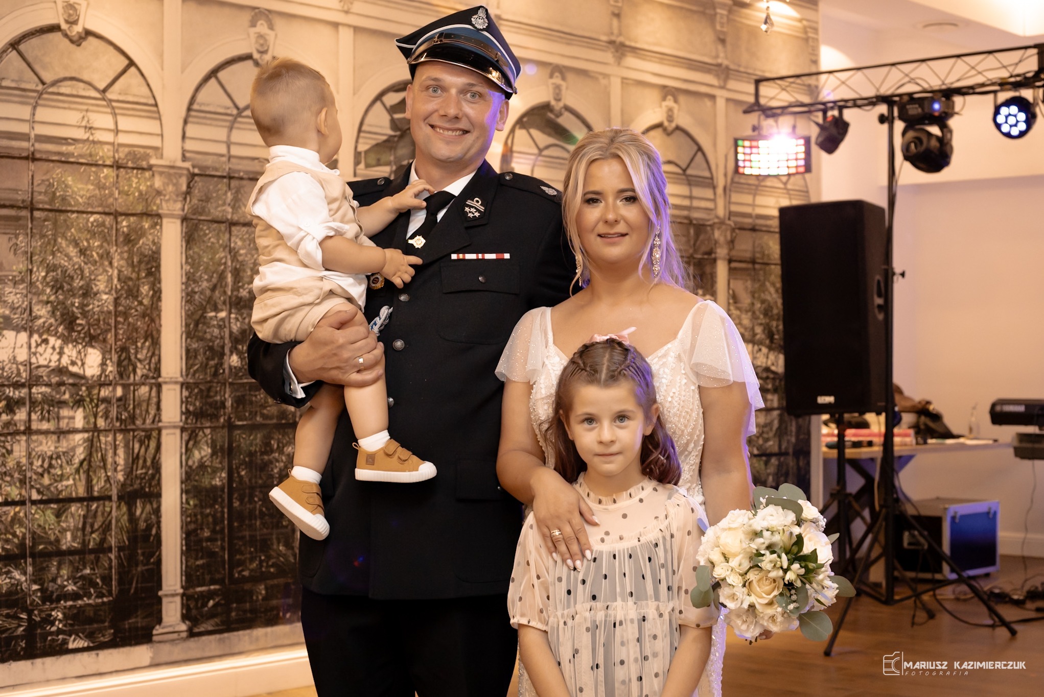 Uśmiechnięta rodzina: mężczyzna w mundurze trzyma synka, obok żona i córka z bukietem kwiatów na tle ściany z motywem architektonicznym.