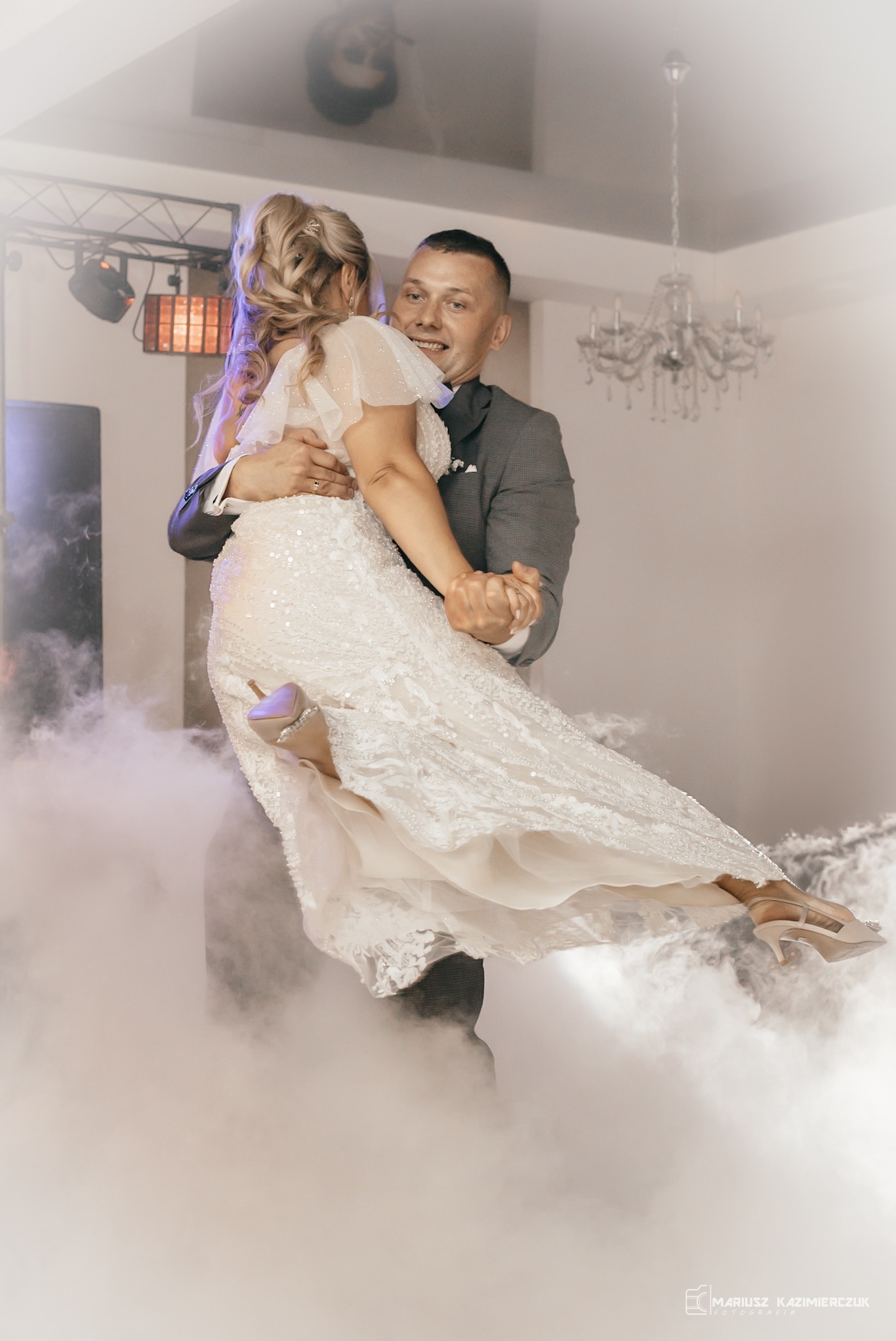 Uśmiechnięty pan młody w szarym garniturze podnosi pannę młodą w białej, błyszczącej sukni podczas pierwszego tańca w dymie scenicznym, widoczny żyrandol i reflektory.