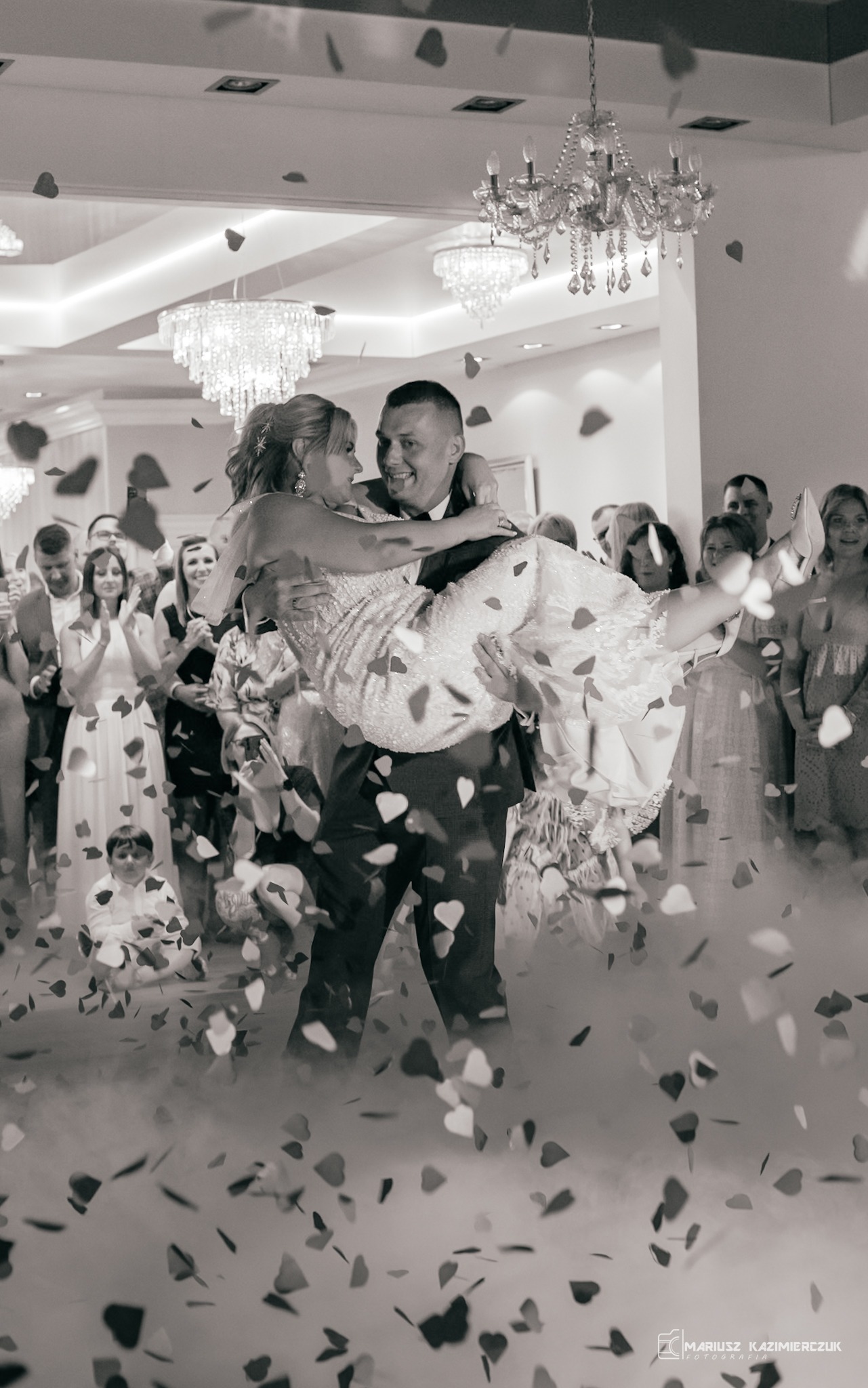 Czarno-białe zdjęcie pana niosącego pannę młodą na rękach podczas pierwszego tańca, z konfetti w kształcie serc unoszącym się w powietrzu, na tle zgromadzonych gości i kryształowych żyrandoli.