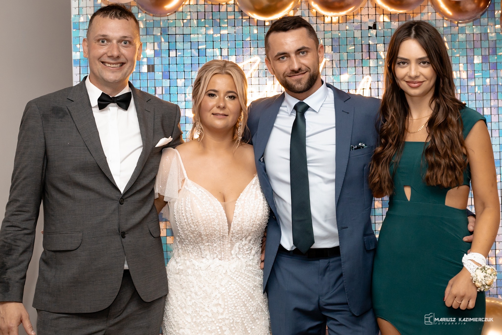 Cztery osoby pozują do zdjęcia na tle błyszczącej ściany: mężczyzna w szarym garniturze z muchą, kobieta w białej sukni z koronki, mężczyzna w niebieskim garniturze z krawatem i kobieta w zielonej...