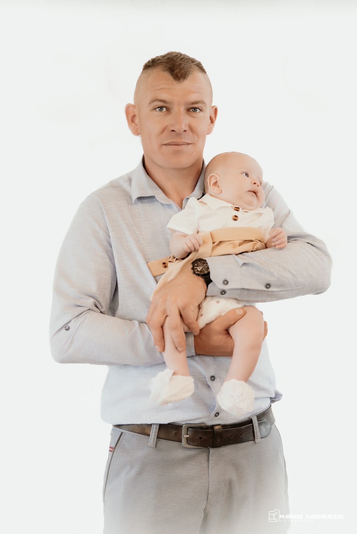 Ojciec z charakterystyczną fryzurą trzymający na rękach niemowlę ubrane w białe ubranko z beżowym pasem, na jasnym tle.