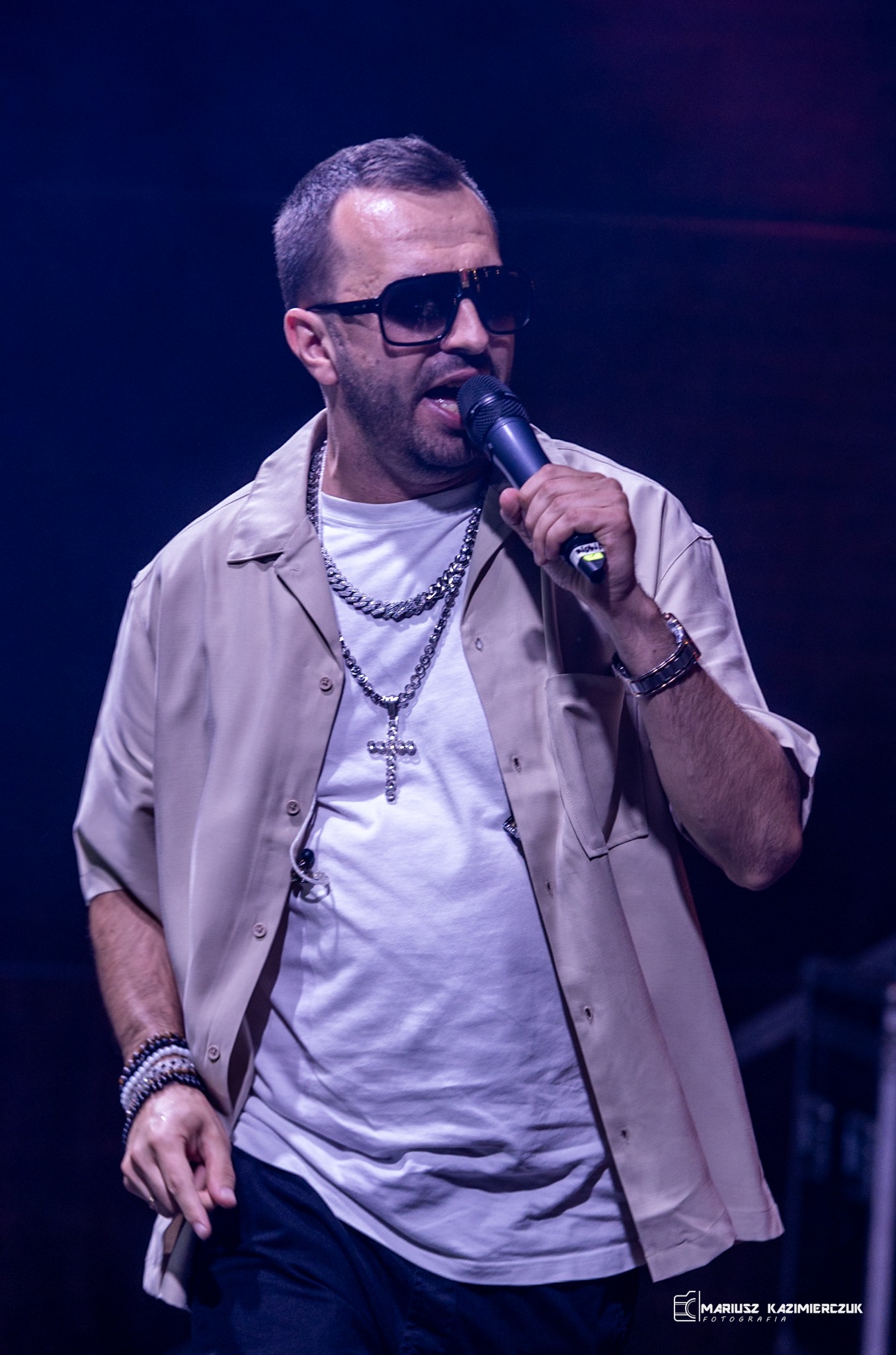 Mężczyzna w okularach przeciwsłonecznych śpiewa do mikrofonu, ubrany w białą koszulkę, beżową koszulę i srebrny łańcuch z krzyżem, na ciemnym tle.