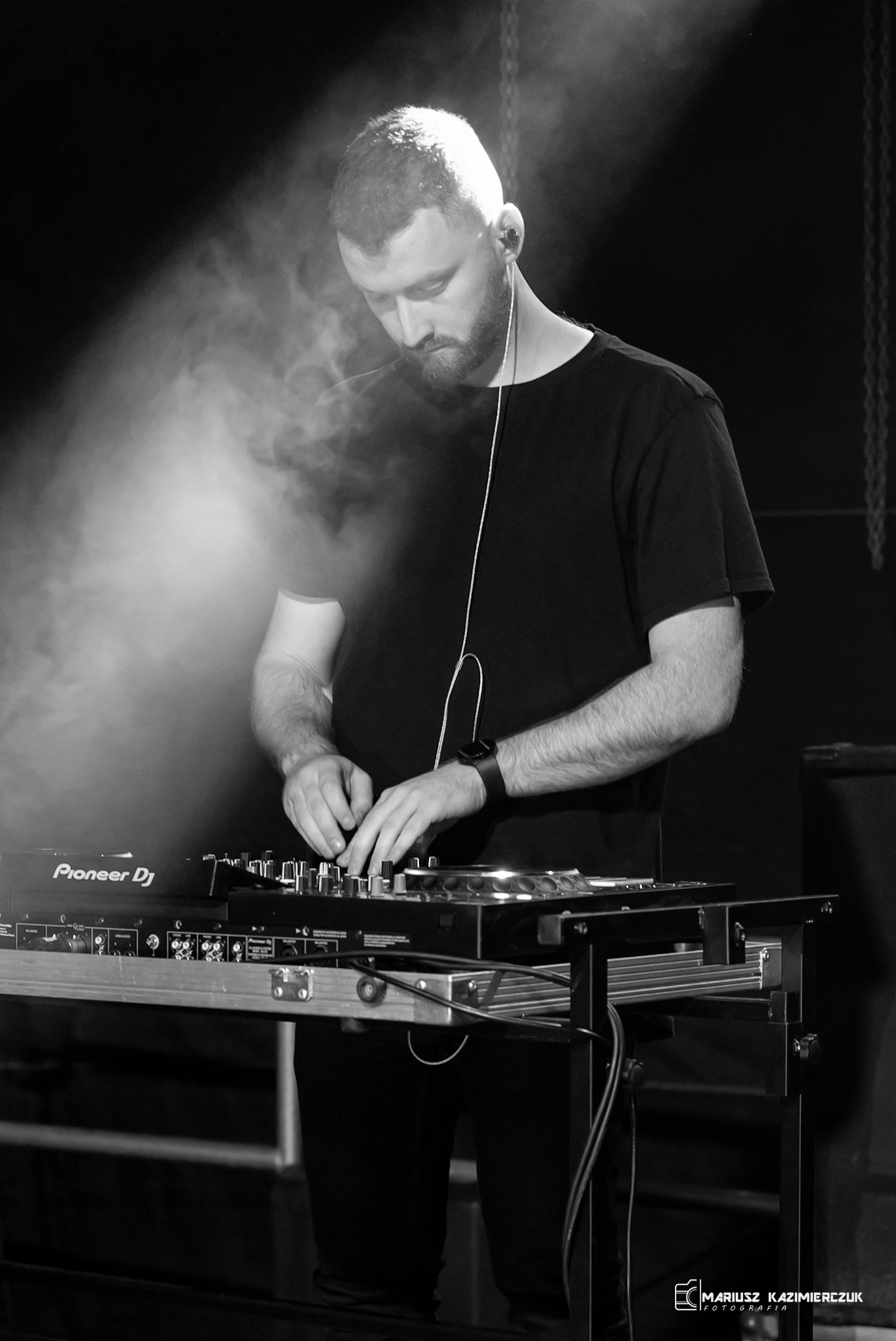Czarno-białe ujęcie DJ-a w trakcie występu, skupionego na miksowaniu muzyki na sprzęcie Pioneer DJ, z delikatną poświatą w tle i widocznymi kablami.