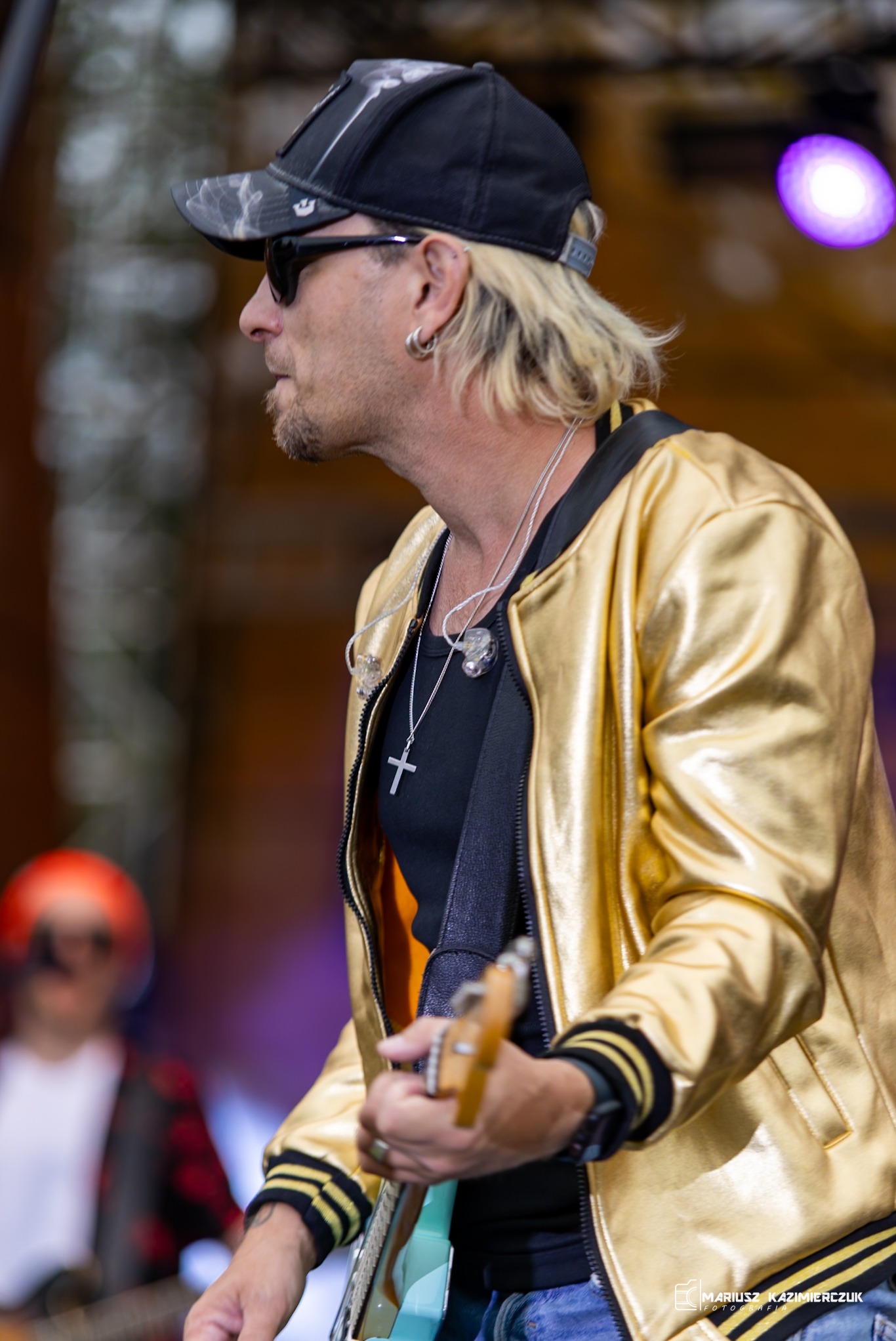 Profil muzyka w czapce i złotej kurtce grającego na gitarze podczas koncertu w Ornecie, widoczny krzyżyk na szyi i słuchawki douszne.