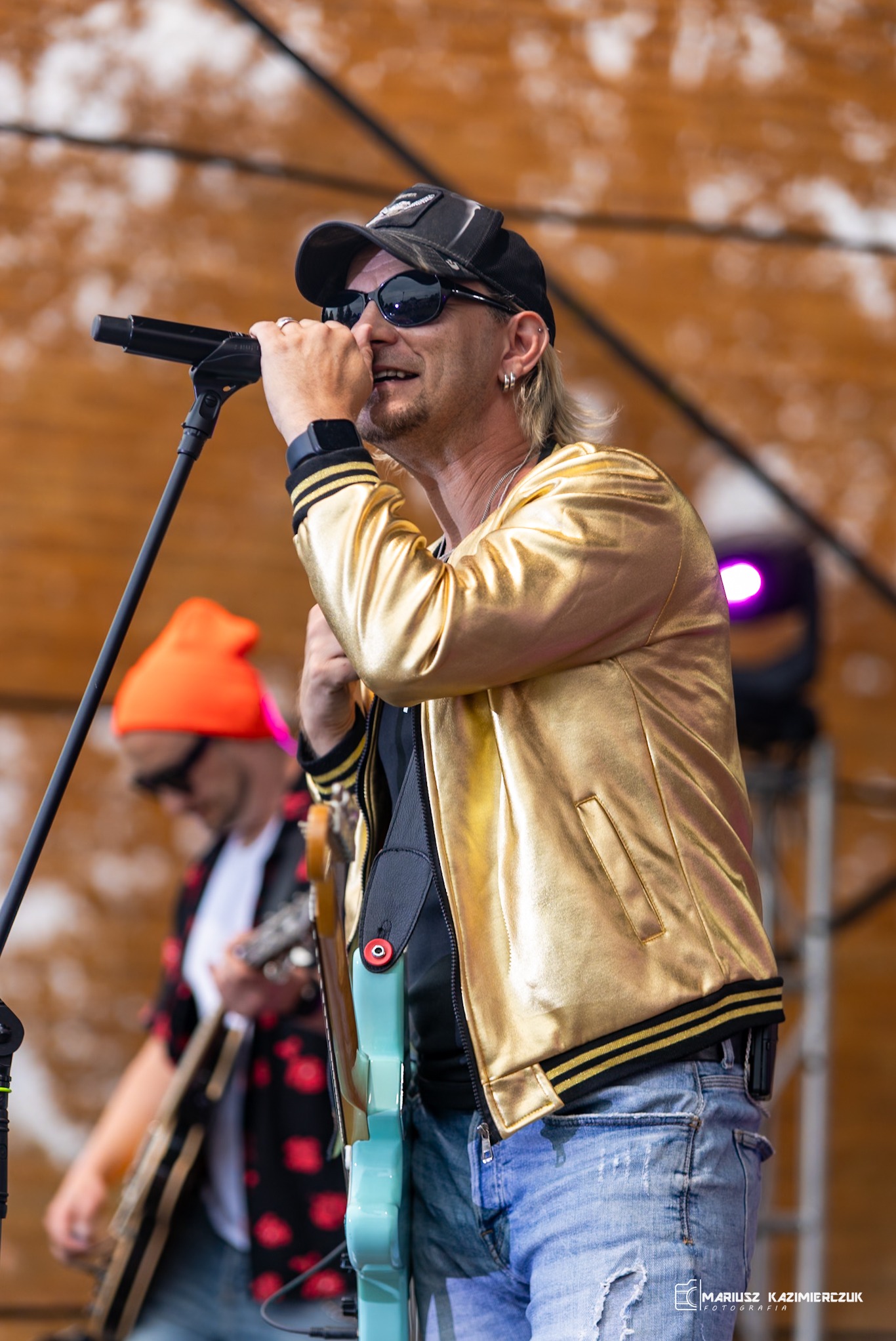 Wokalista w złotej kurtce i czapce śpiewa do mikrofonu na scenie, w tle gitarzysta z pomarańczową czapką.