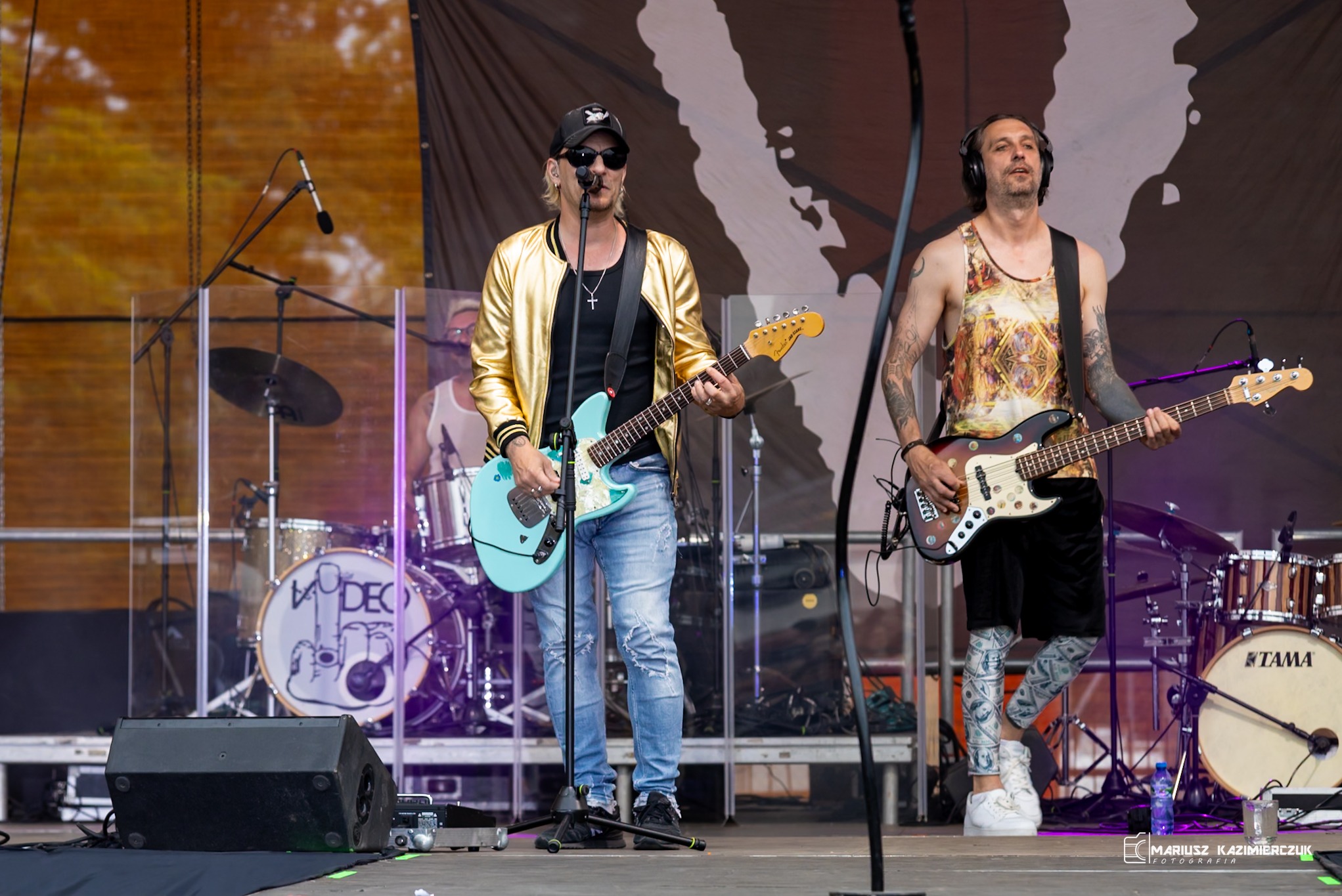 Zespół muzyczny grający na scenie: gitarzysta w złotej kurtce i basista z tatuażami, perkusja w tle.