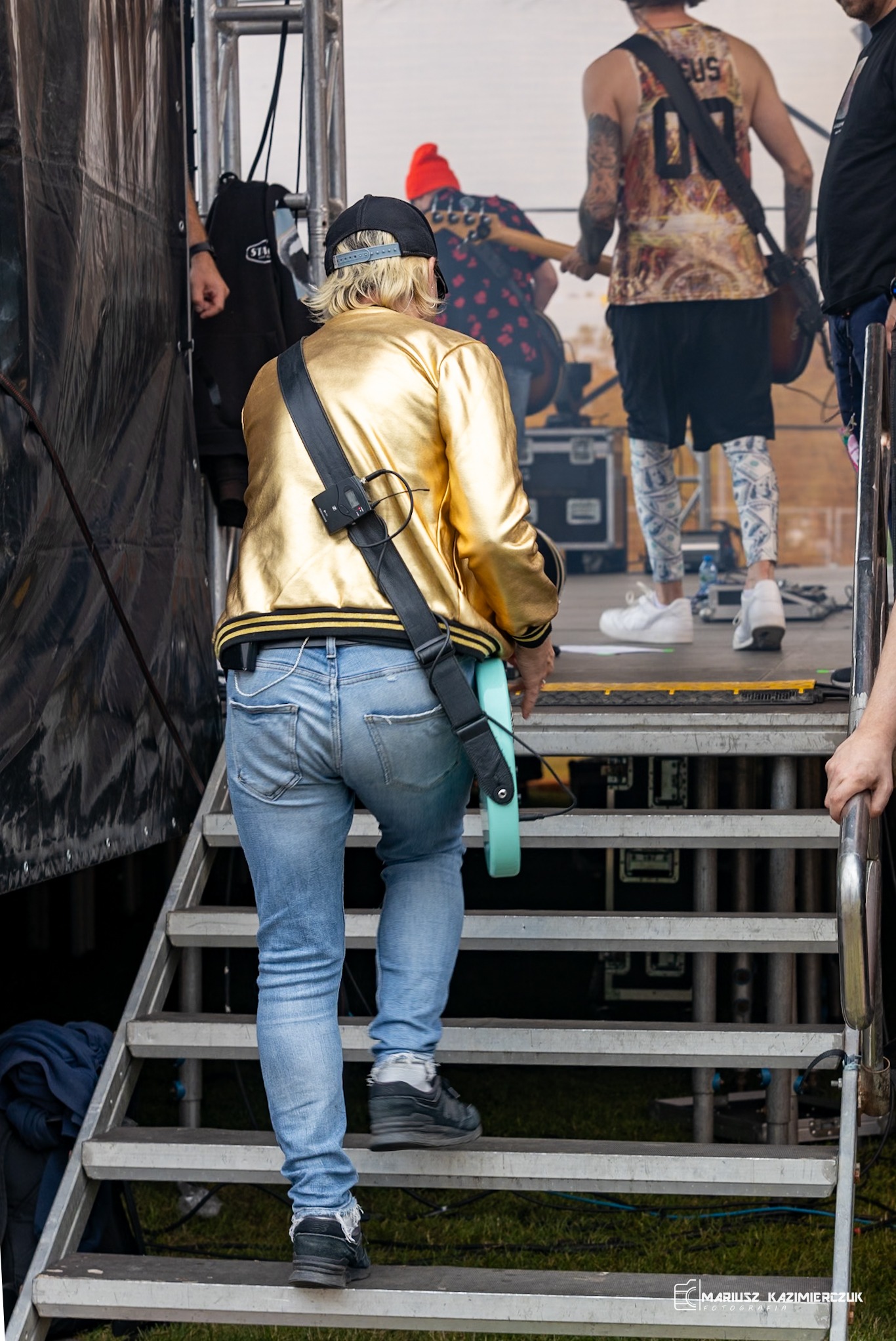 Osoba w złotej kurtce wchodzi po schodach na scenę, trzymając gitarę; widoczni muzycy i sprzęt muzyczny w tle.