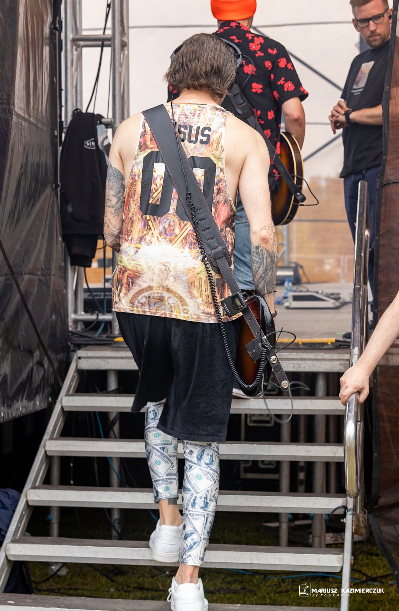 Muzyk z gitarą wchodzi po schodach na scenę, ubrany w koszulkę z nadrukiem i legginsy z motywem banknotów dolarowych.